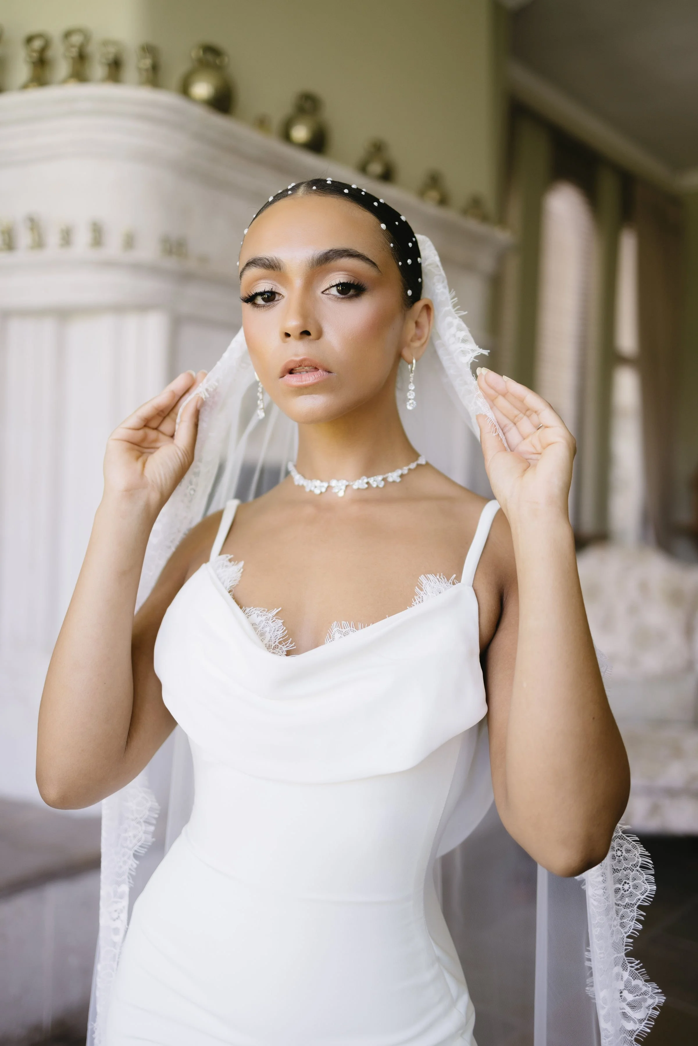 A woman dressed in a white wedding gown with a lace-trimmed veil, holding the veil with both hands, in an elegant indoor setting with a fireplace and decorative vases in the background.