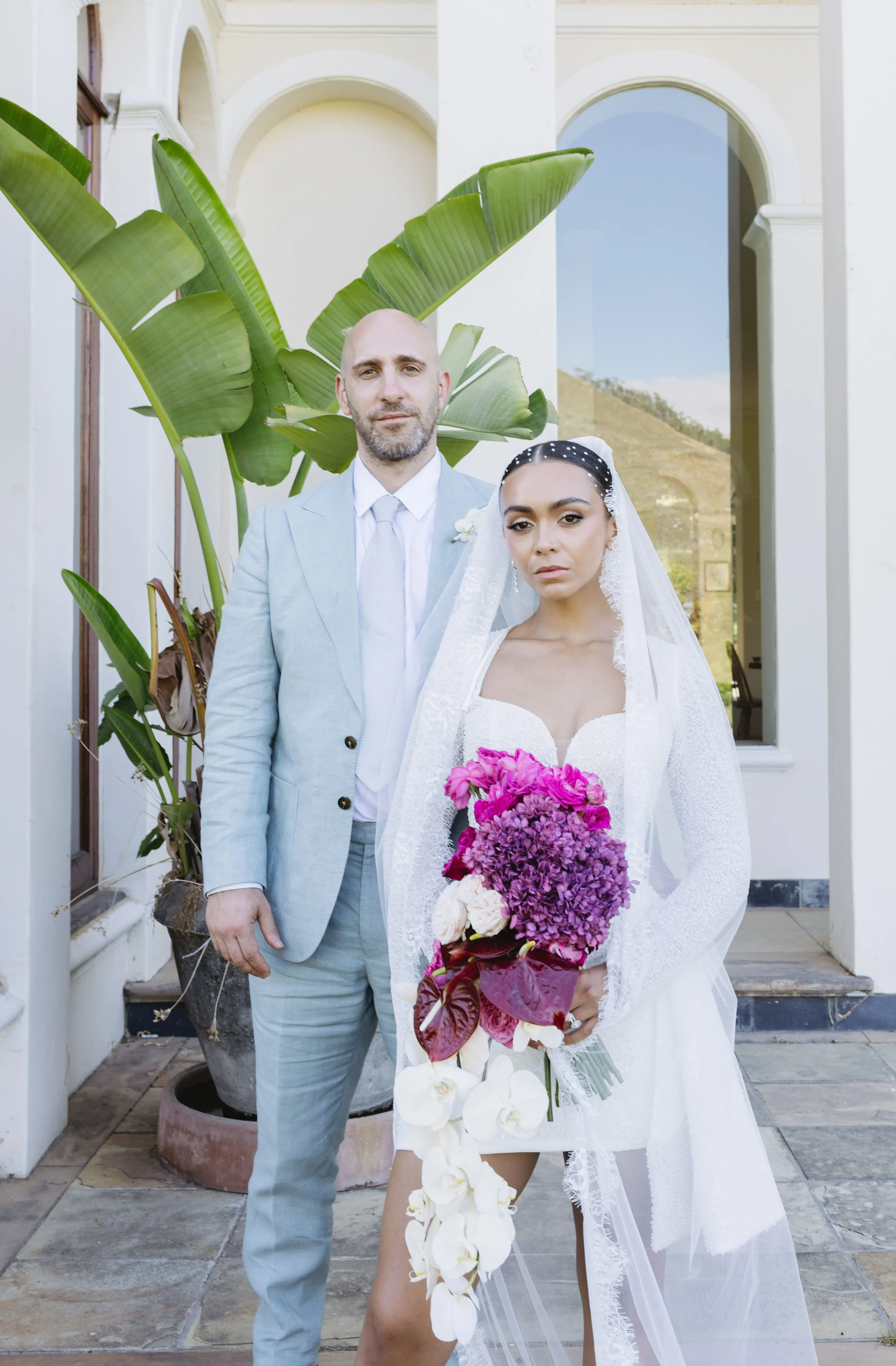 A bride and groom standing outdoors in front of a white building with large windows and tropical plants, with the bride holding a bouquet of pink, purple, and white flowers.