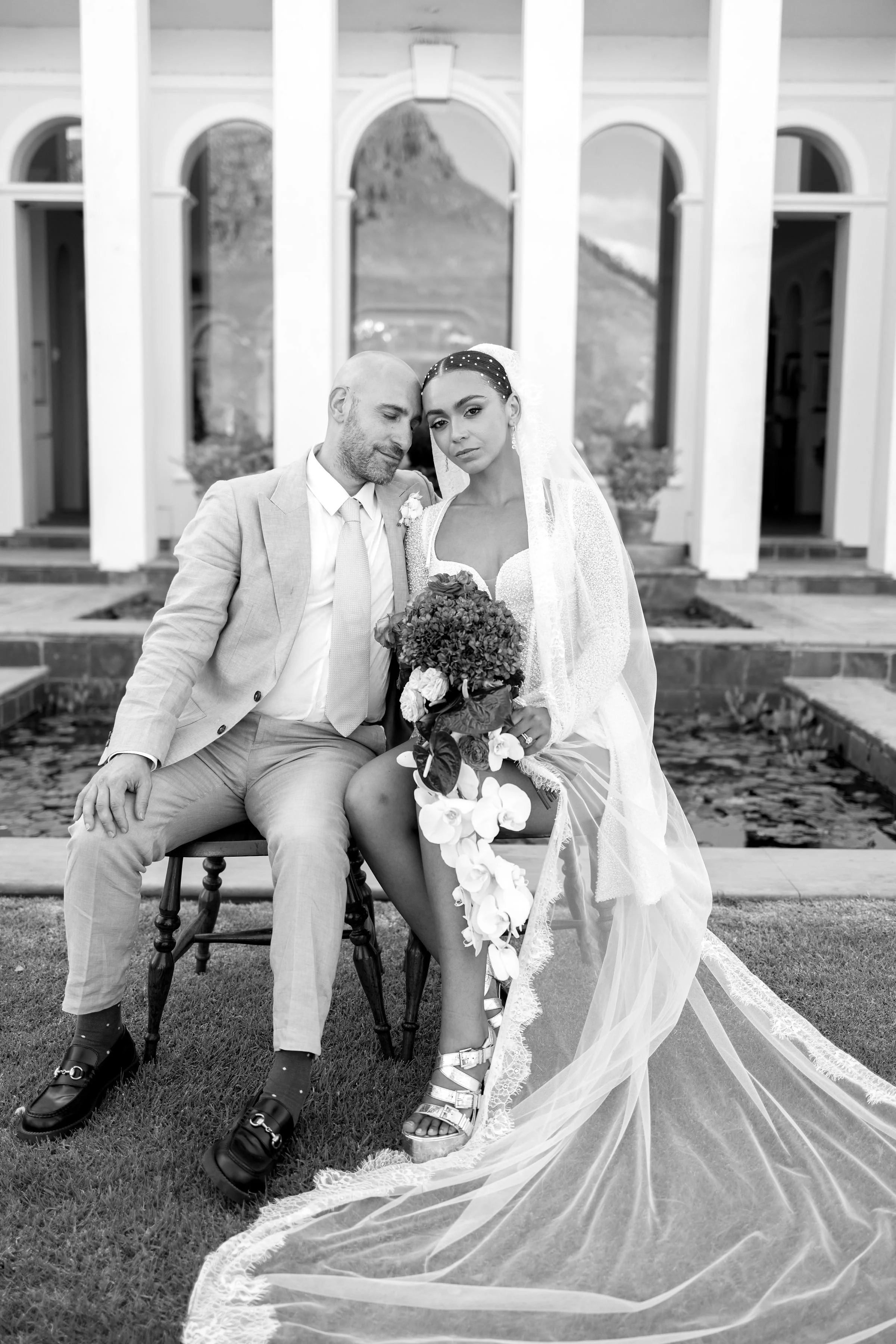 Black and white photo of a bride and groom sitting outside a building with arches, holding a bouquet of flowers, with a mountain in the background.
