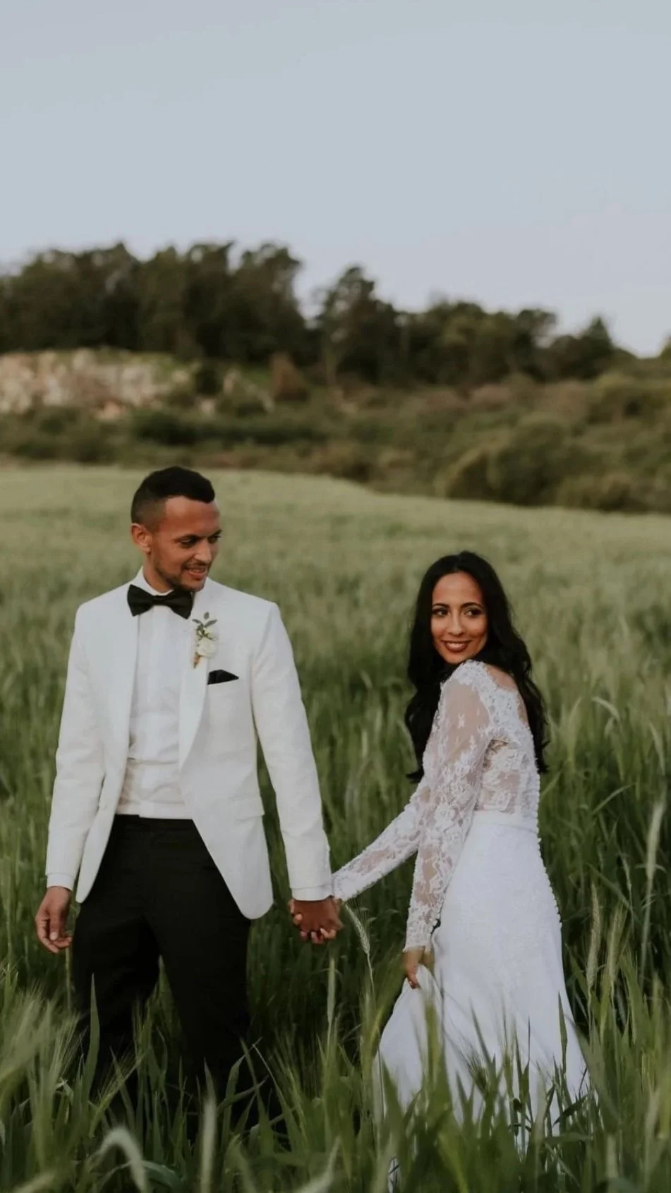 A newlywed couple in wedding attire holding hands and smiling in a green field.