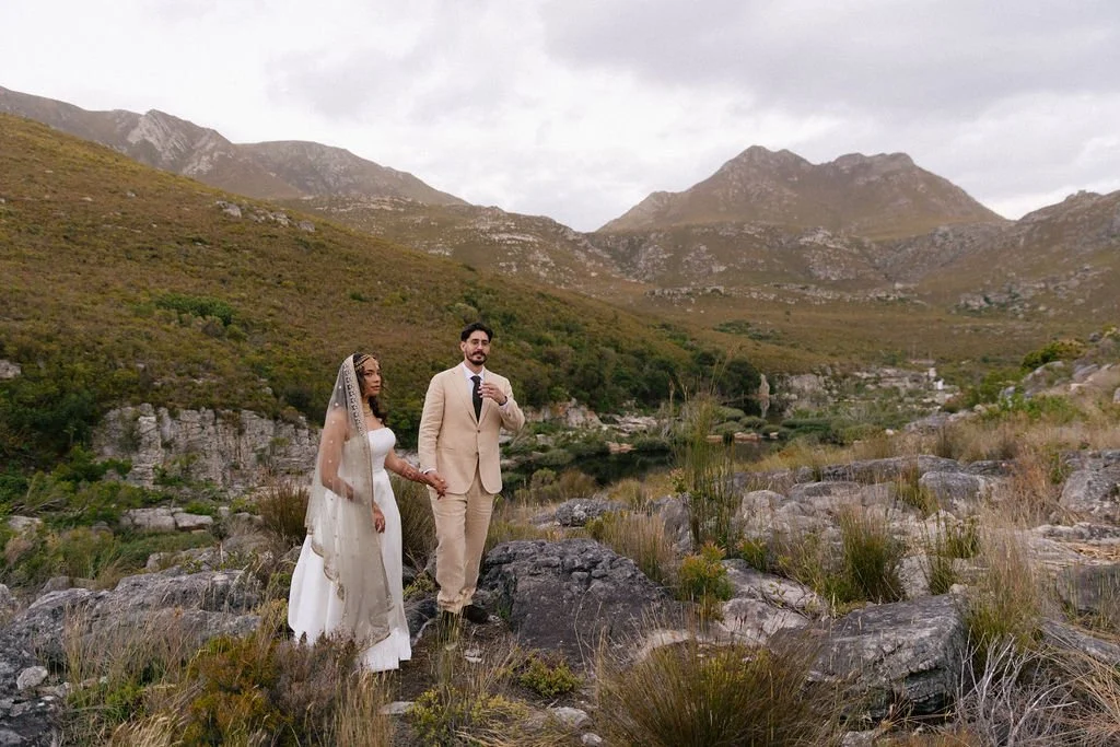 A bride and groom holding hands in a mountainous outdoor setting, with rocks and sparse vegetation, under a cloudy sky.