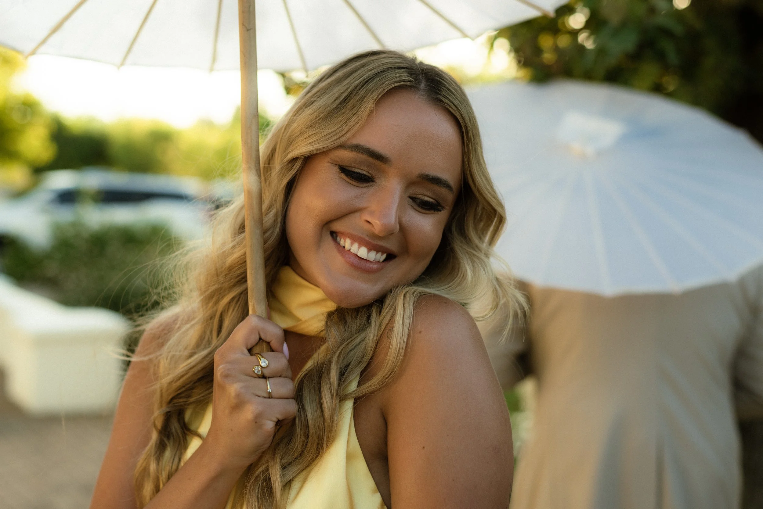 A woman with long blonde hair smiling while holding a white umbrella in an outdoor setting.