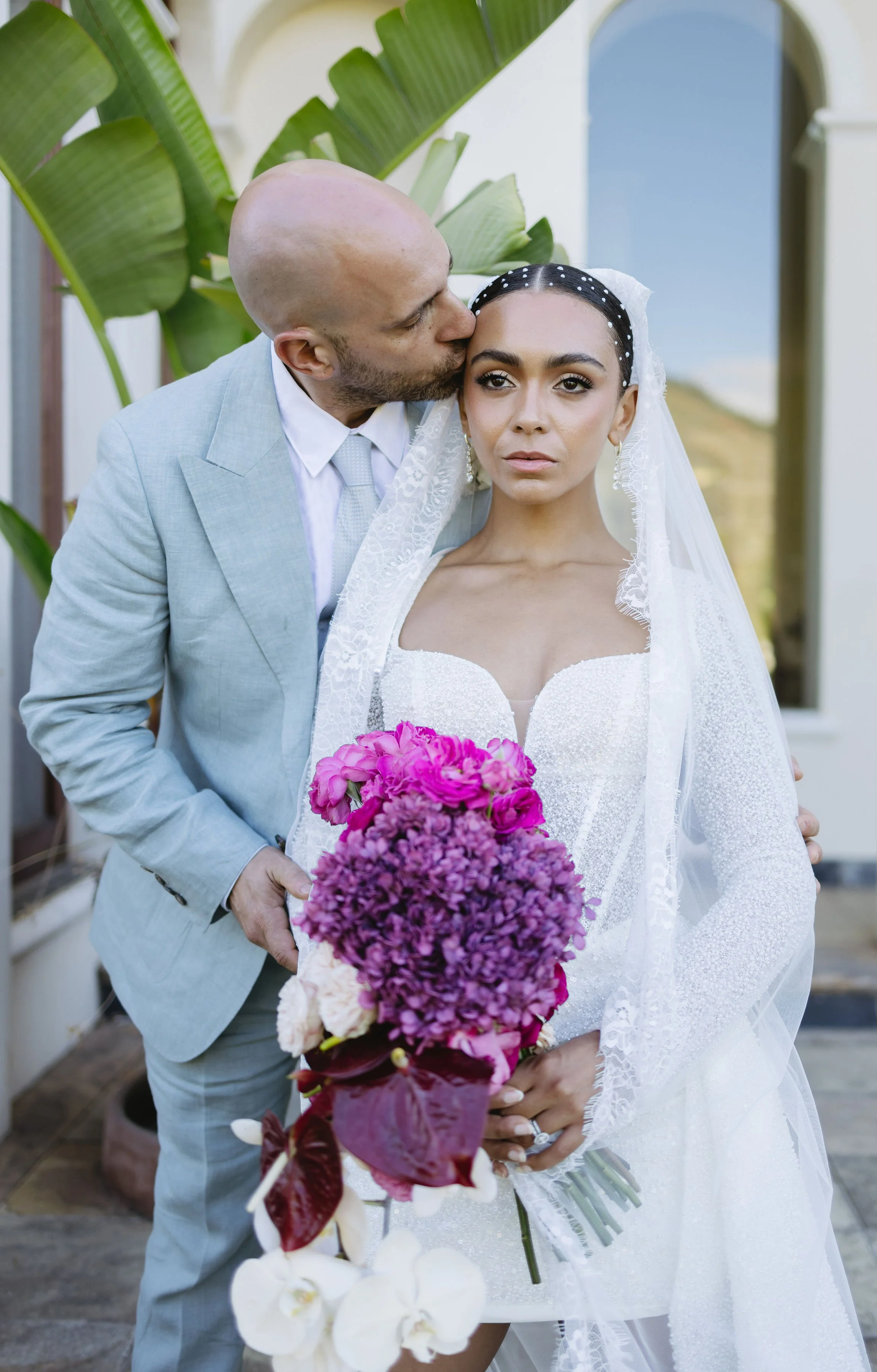 A man in a gray suit kisses a woman in a white wedding dress on her forehead while she holds a colorful bouquet of pink and purple flowers.