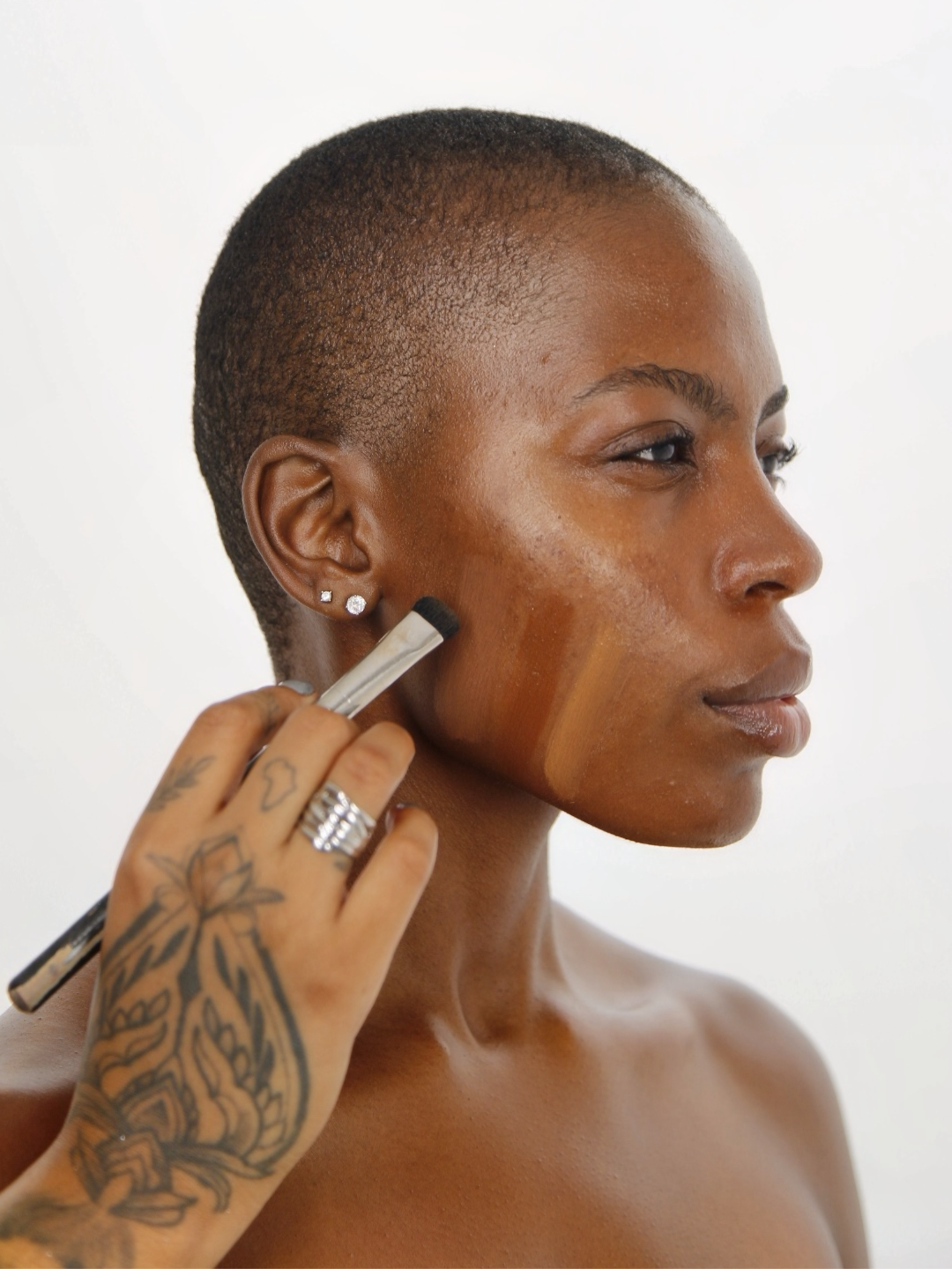 A woman with short hair receiving makeup application on her cheek with a brush by an artist with a tattooed hand and earrings.