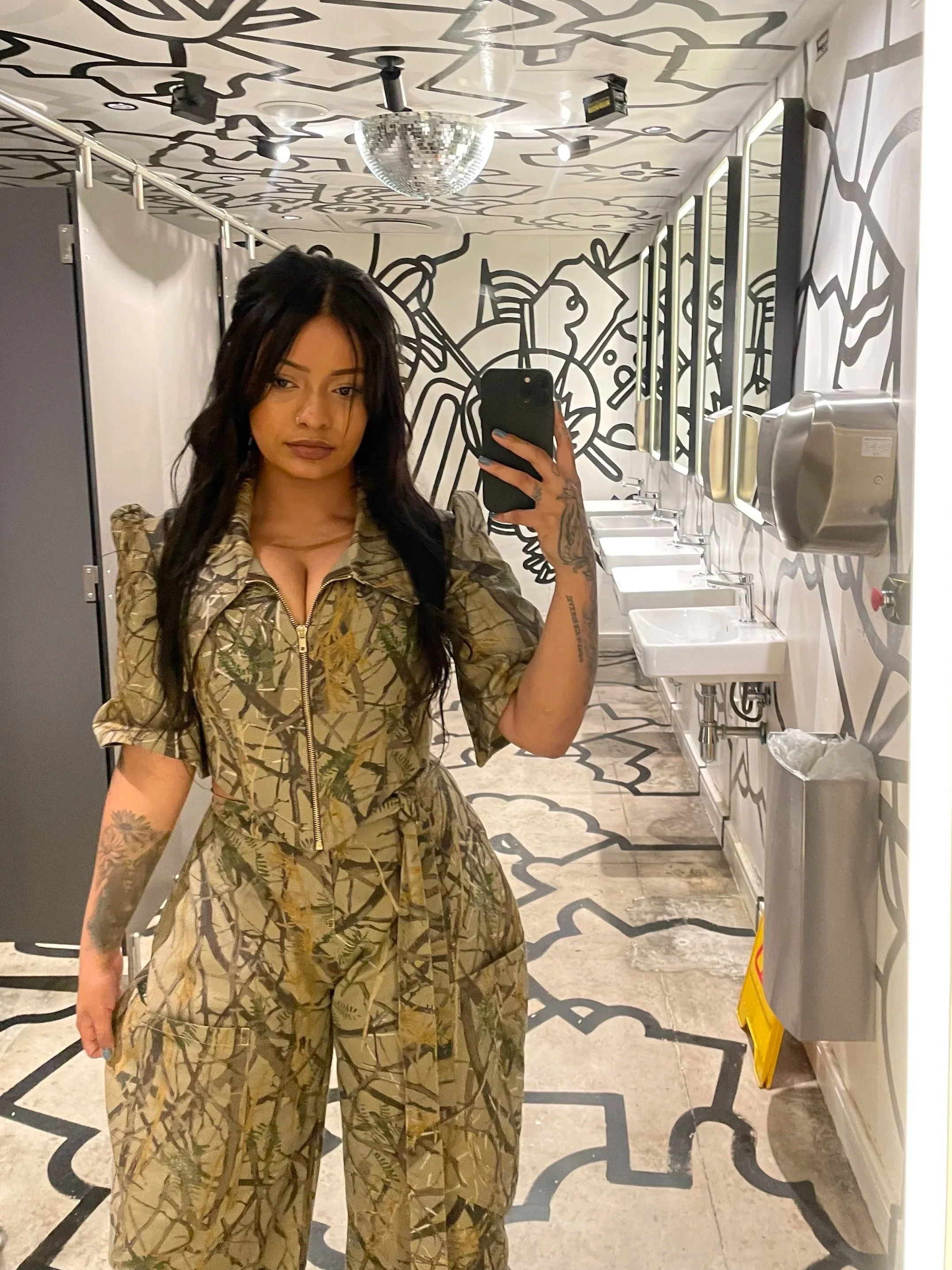 A woman taking a mirror selfie in a bathroom with black and white abstract wall and ceiling art, featuring a disco ball light fixture, sinks, and a soap dispenser.