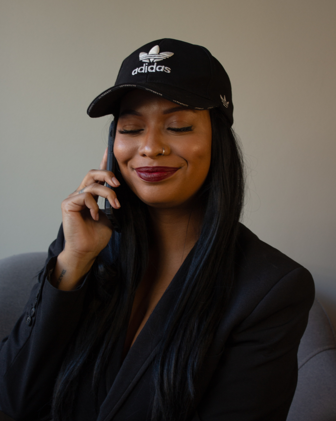 A woman with long black hair wearing a black Adidas cap and black blazer, talking on a phone with a slight smile and closed eyes.