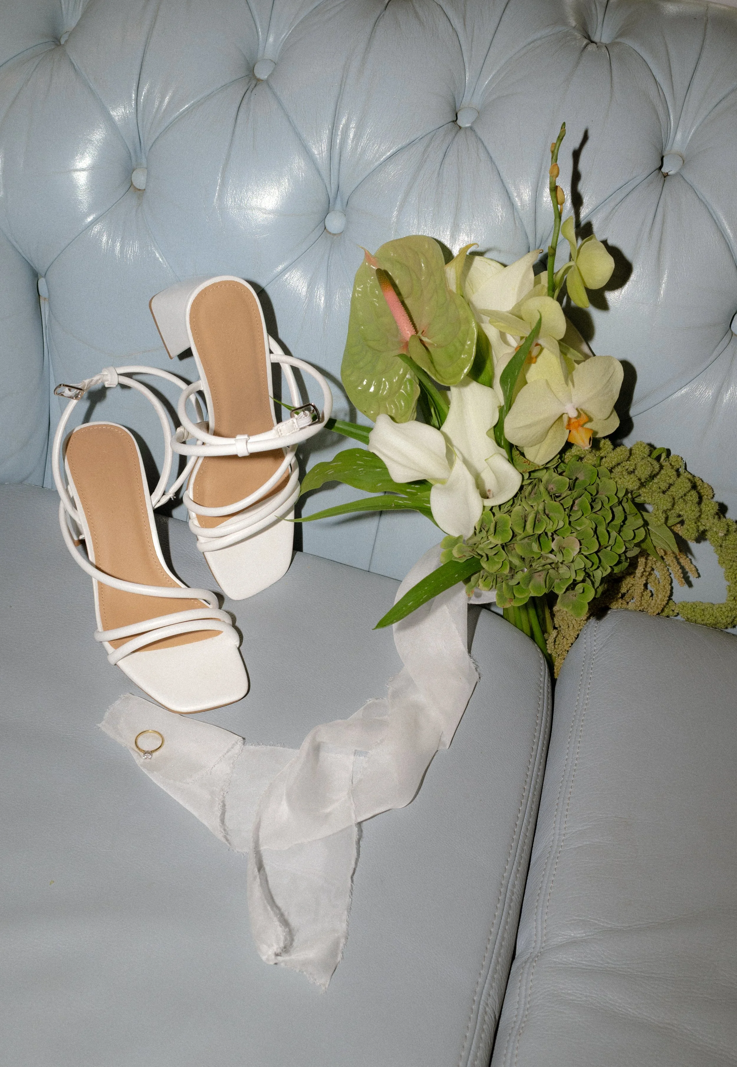 A pair of white high-heeled sandals, a bouquet of flowers, and a gold ring on a ribbon, placed on a light blue tufted leather sofa.