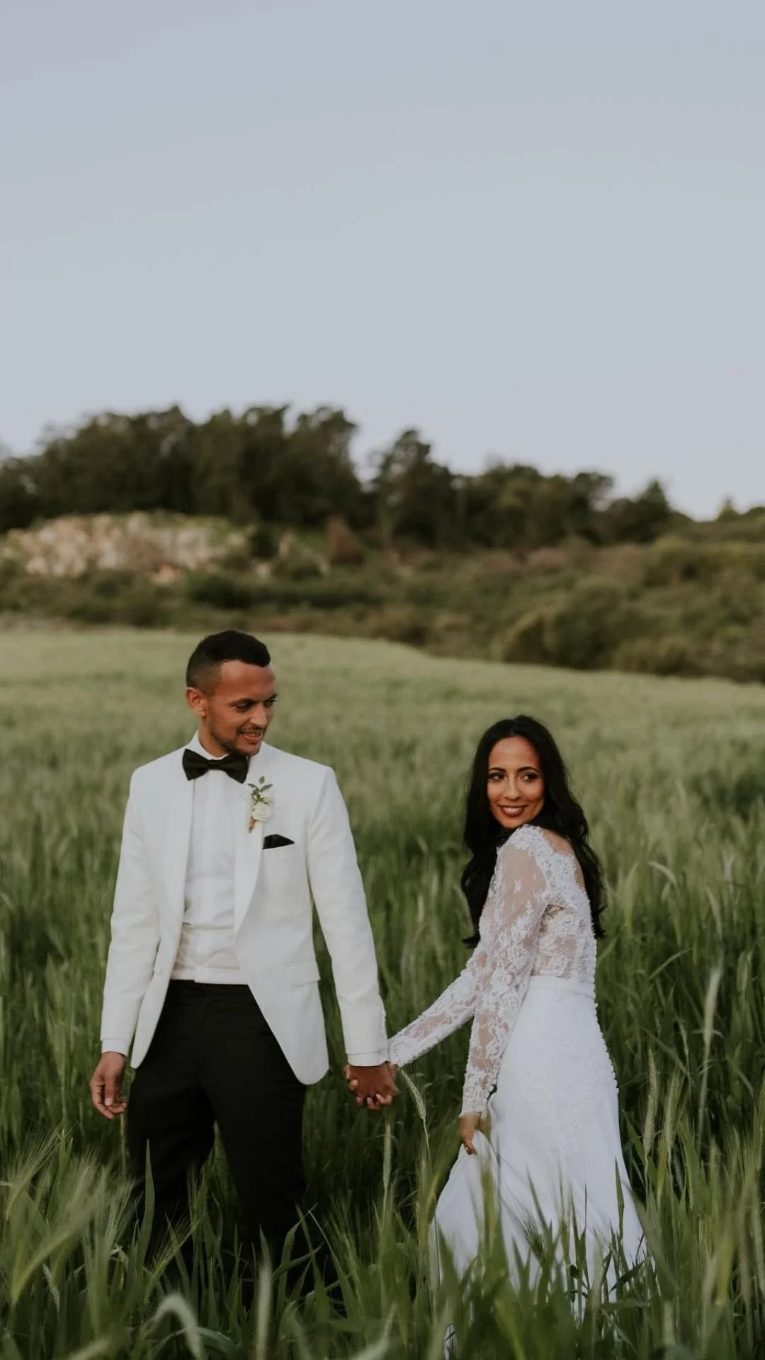 A groom and bride holding hands in a green field, smiling at each other. The groom is wearing a white tuxedo with black bow tie and pants, and the bride is in a white lace wedding dress with long sleeves.