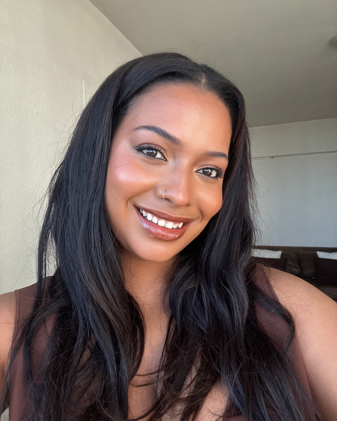 A young woman with long black wavy hair, smiling, wearing a sleeveless brown top, in a room with a white wall and a brown couch in the background.