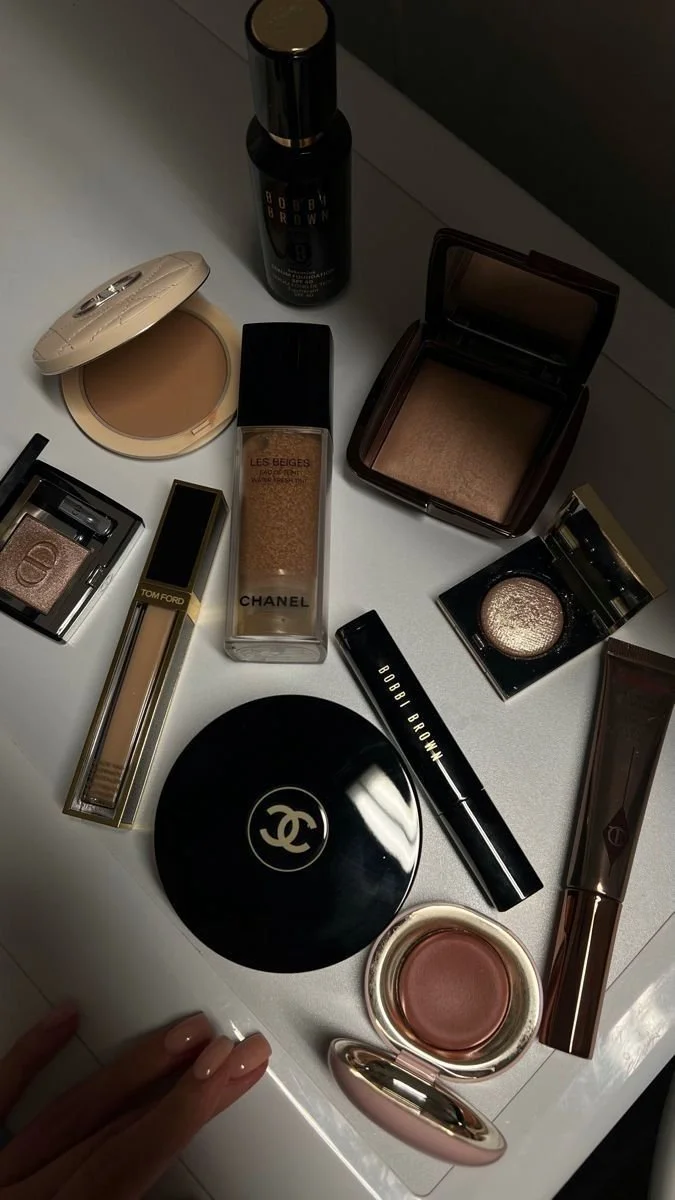 A collection of makeup and beauty products on a white surface, including powders, a foundation spray, highlighters, and a compact from Chanel, Bobbi Brown, Tom Ford, and Dior.