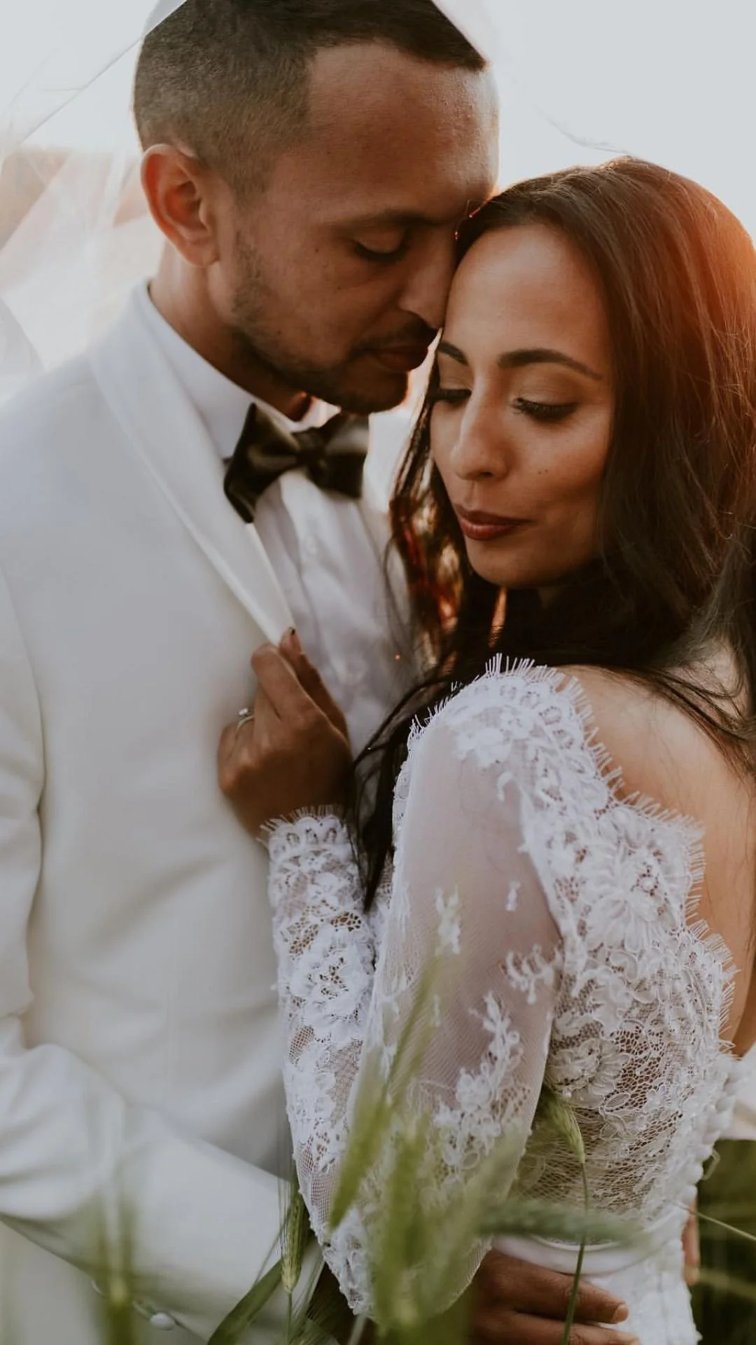 A groom in a white suit and black bow tie with a bride in a white lace dress sharing an intimate moment outdoors during sunset, with their foreheads touching and eyes closed.