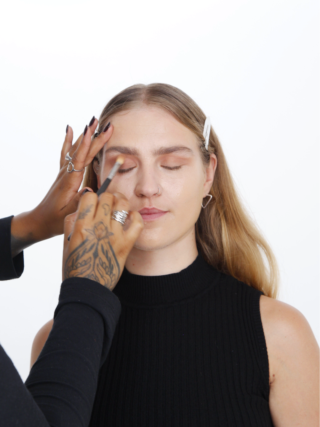 A woman with long light brown hair getting makeup applied to her eyelid by a makeup artist with tattoos on their hand, wearing a black long-sleeve shirt, against a white background.