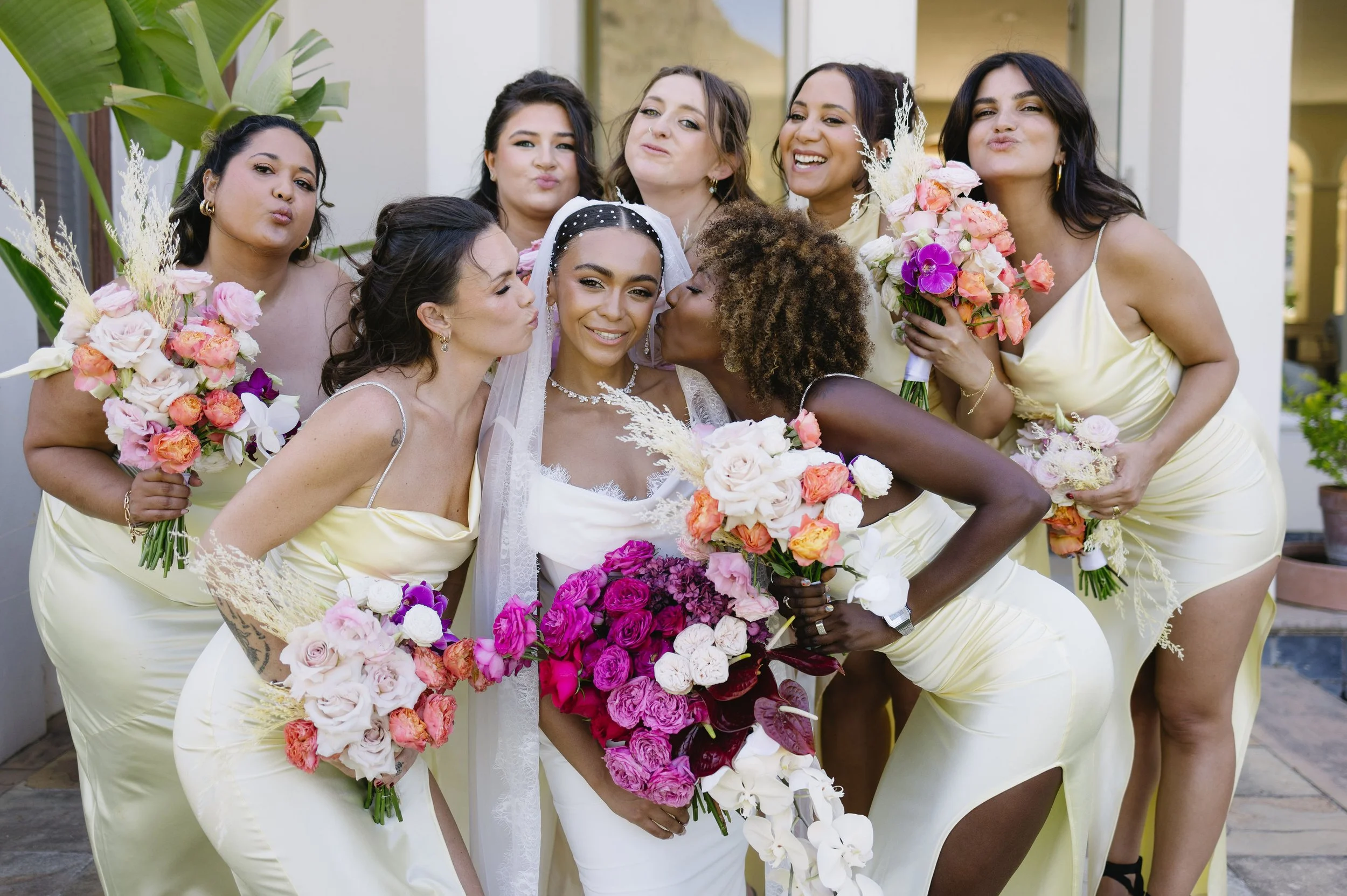 A bride in a white wedding dress and veil surrounded by seven women in pale yellow dresses, all holding bouquets of pink, purple, and white flowers. The women are kissing and posing for a photo at a wedding.