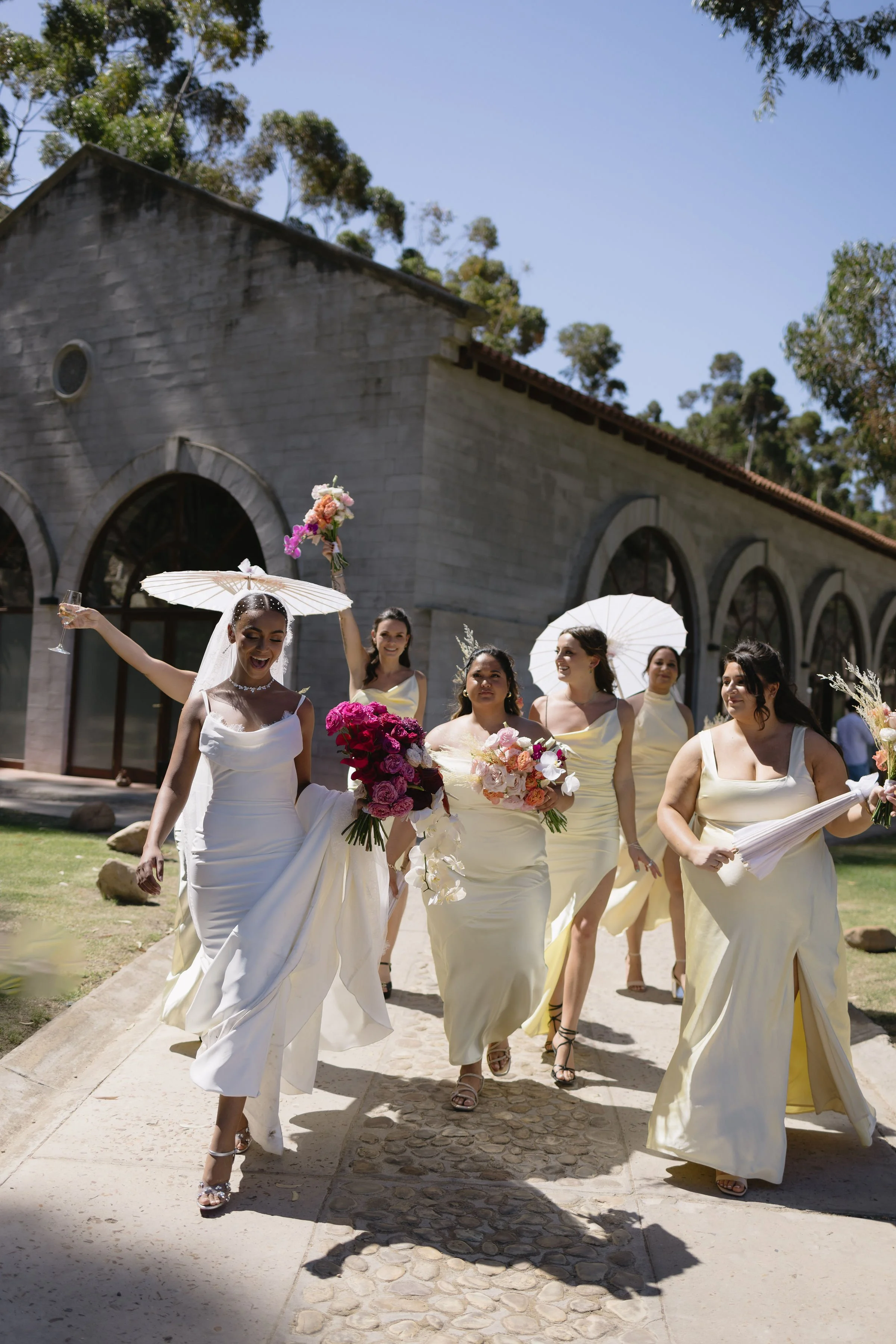 Bridal party walking outdoors on a sunny day, holding umbrellas and bouquets, with a stone building and trees in the background.