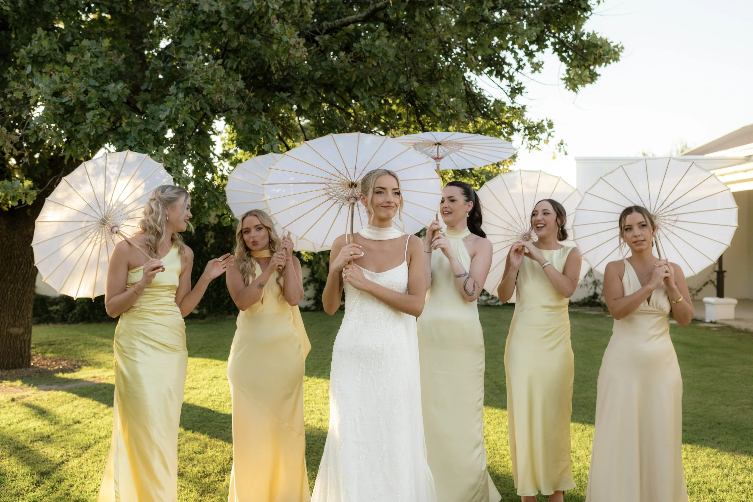 A bride in a white wedding dress standing outdoors with five bridesmaids dressed in yellow gowns, holding white parasols under the shade of a large tree on a sunny day.