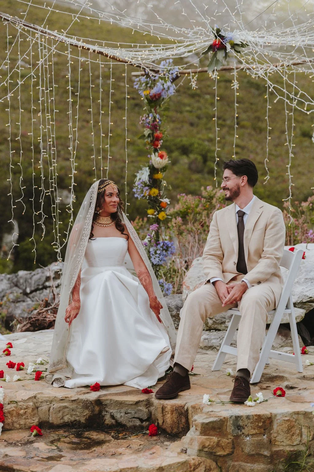 A bride and groom sitting outdoors during their wedding ceremony, decorated with string lights and flowers, with a natural landscape in the background.