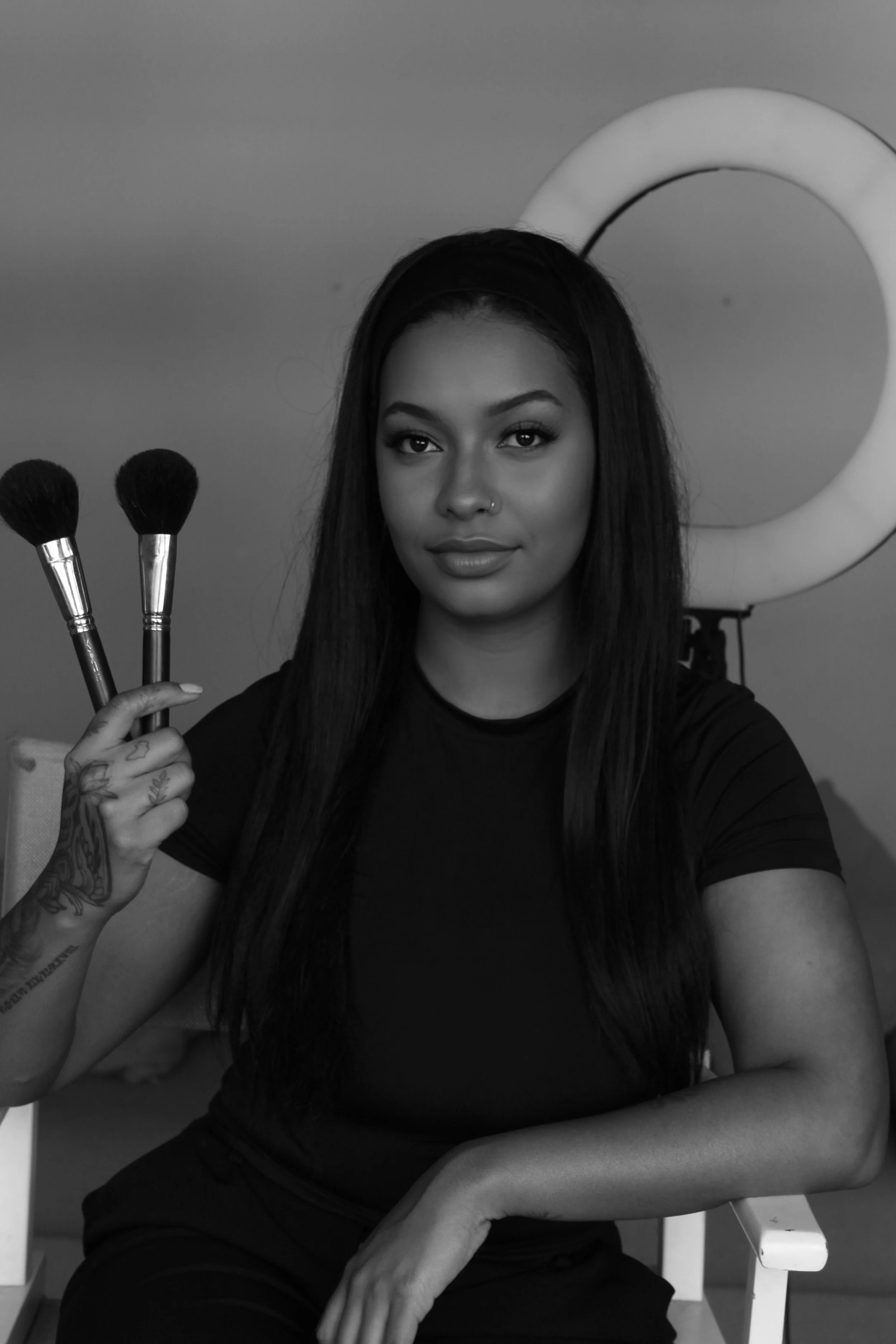 Black and white portrait of a woman with long dark hair, holding two makeup brushes, sitting in front of a ring light.
