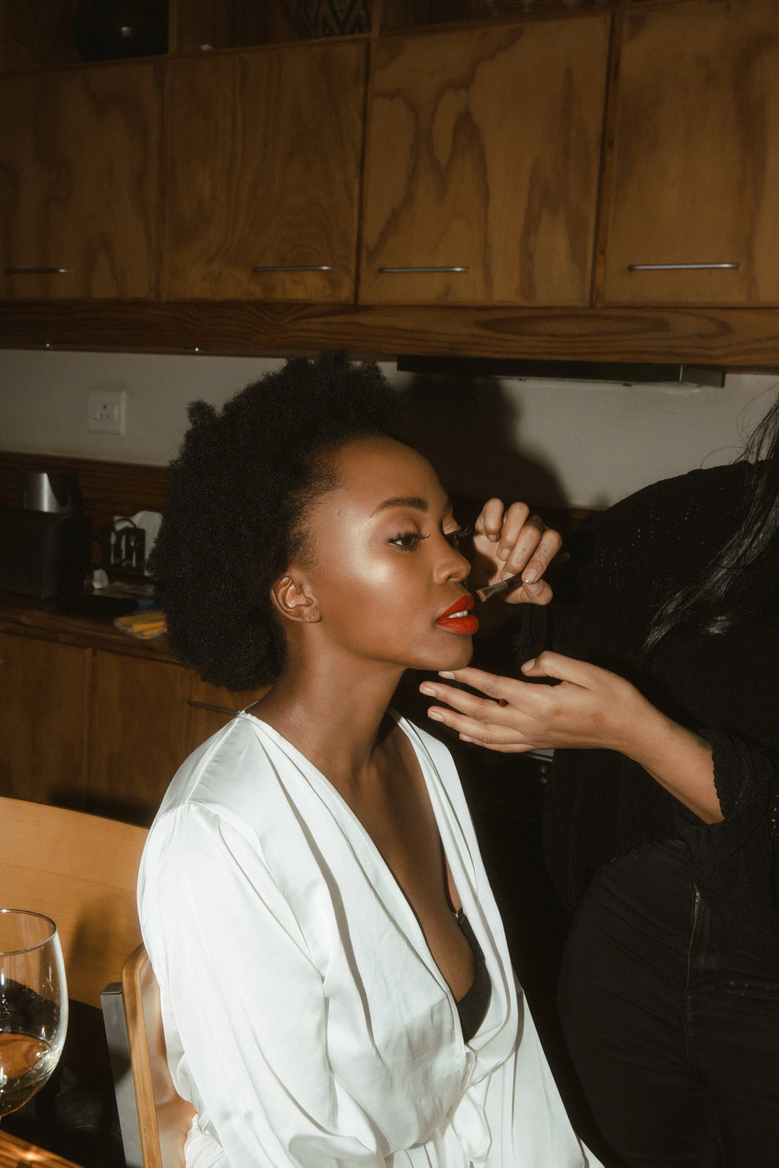 A woman with short curly hair and red lipstick is having makeup applied by a makeup artist in a room with wooden cabinets.