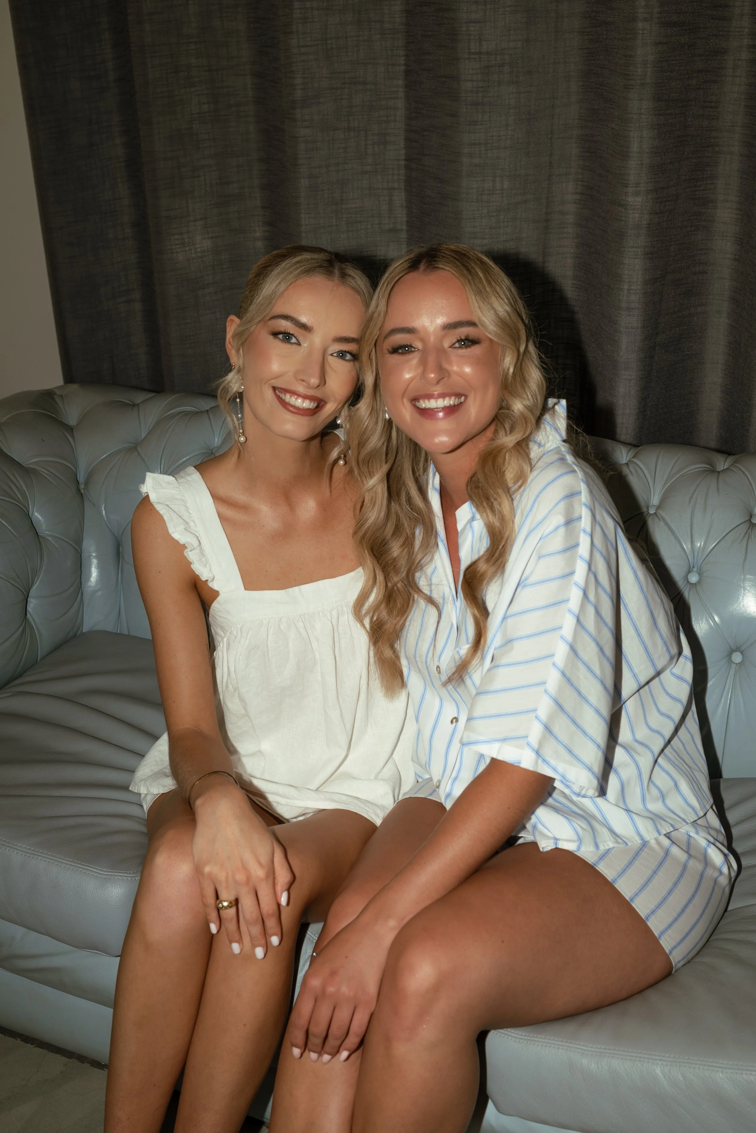 Two women smiling and sitting close together on a grey tufted couch in front of dark curtains, one wearing a white sleeveless dress with ruffles and the other in a blue and white striped shirt and shorts.