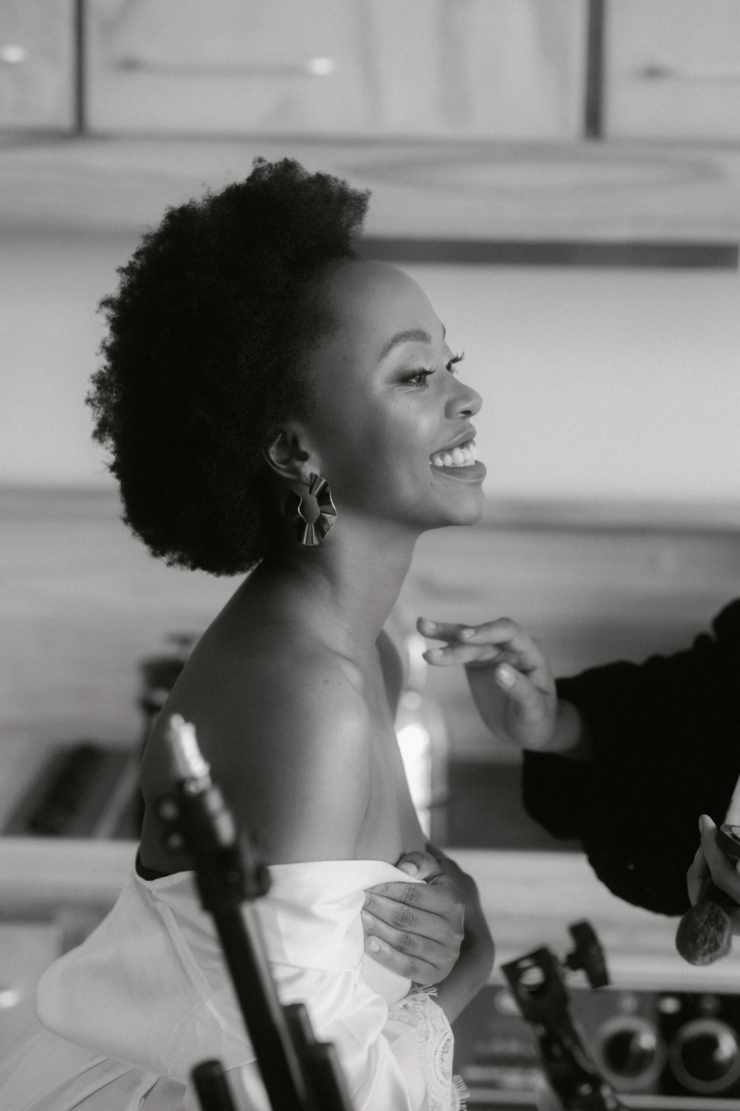 A woman with natural hair, smiling and looking to the side, with a makeup artist touching her shoulder. The scene is in a well-lit room, possibly a studio or dressing area.