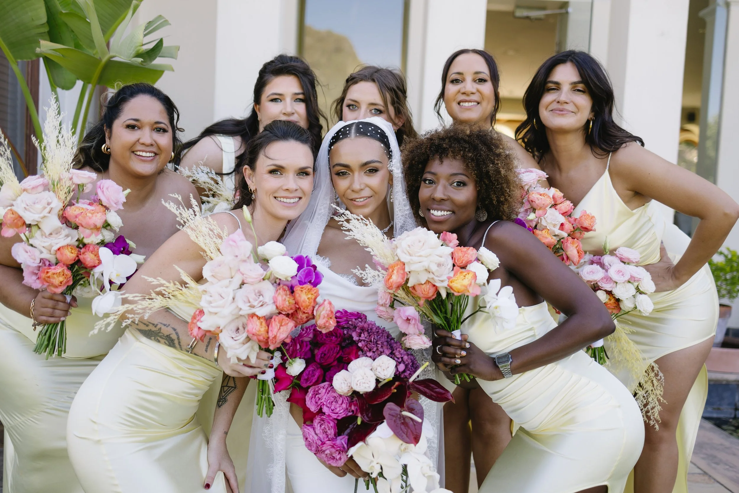 Group of women, including a bride in a white dress and veil, holding colorful bouquets of flowers, smiling outdoors in front of a building with glass windows.