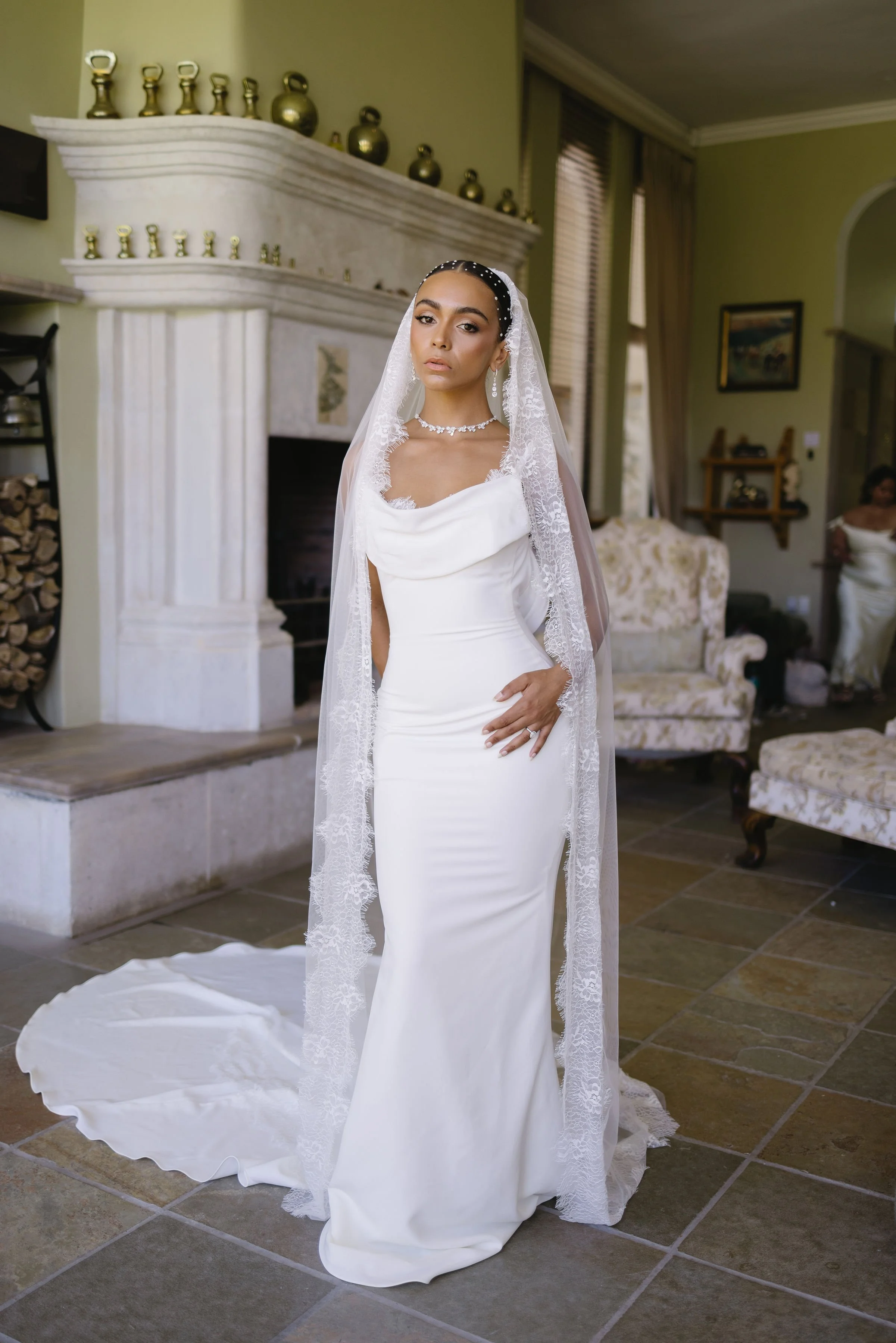 A bride in a white wedding dress with a lace veil standing in a living room with a fireplace, wood mantel, and furniture, looking directly at the camera.