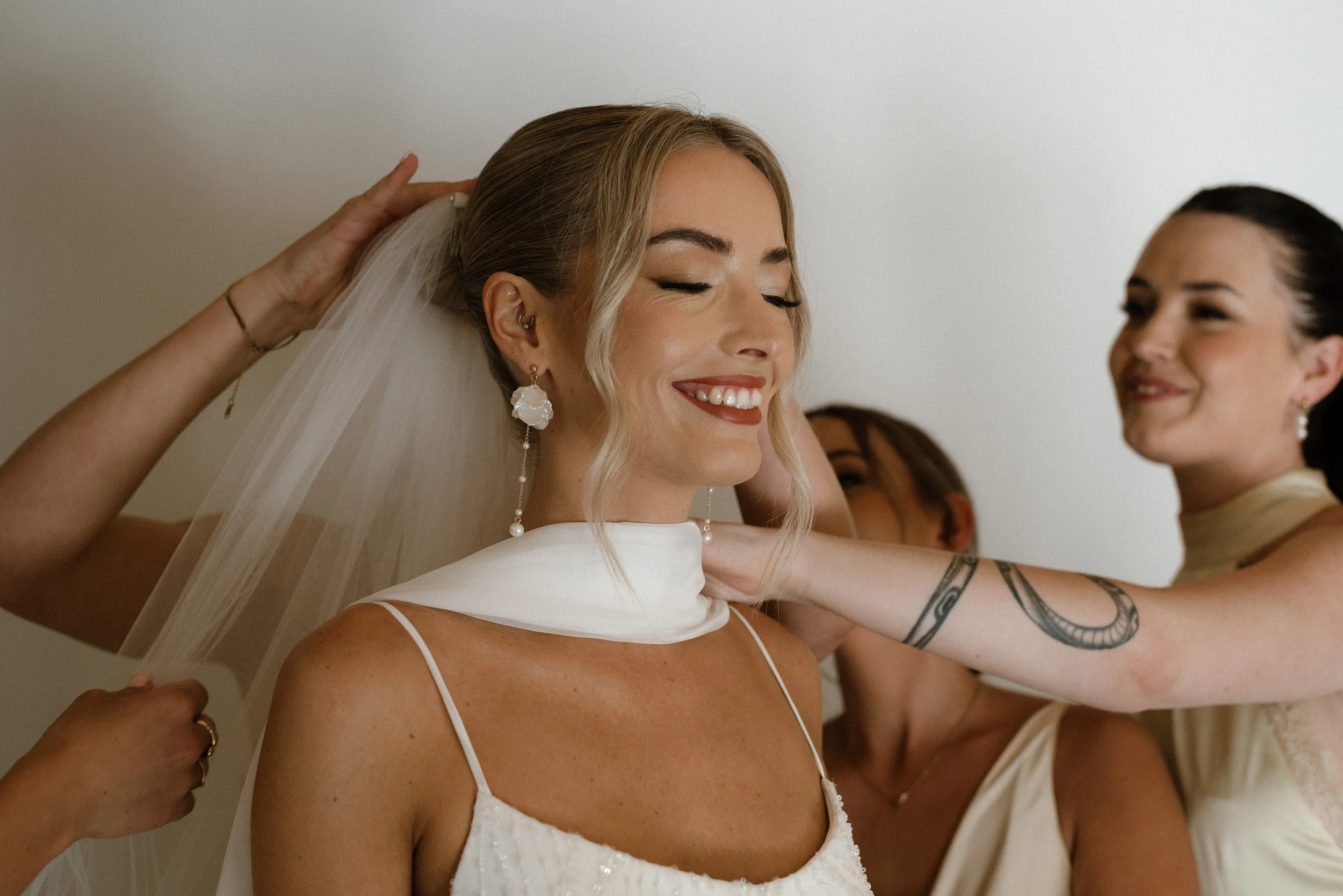 A bride getting ready with help from friends or family, smiling with closed eyes, wearing a white dress, veil, and pearl earrings.