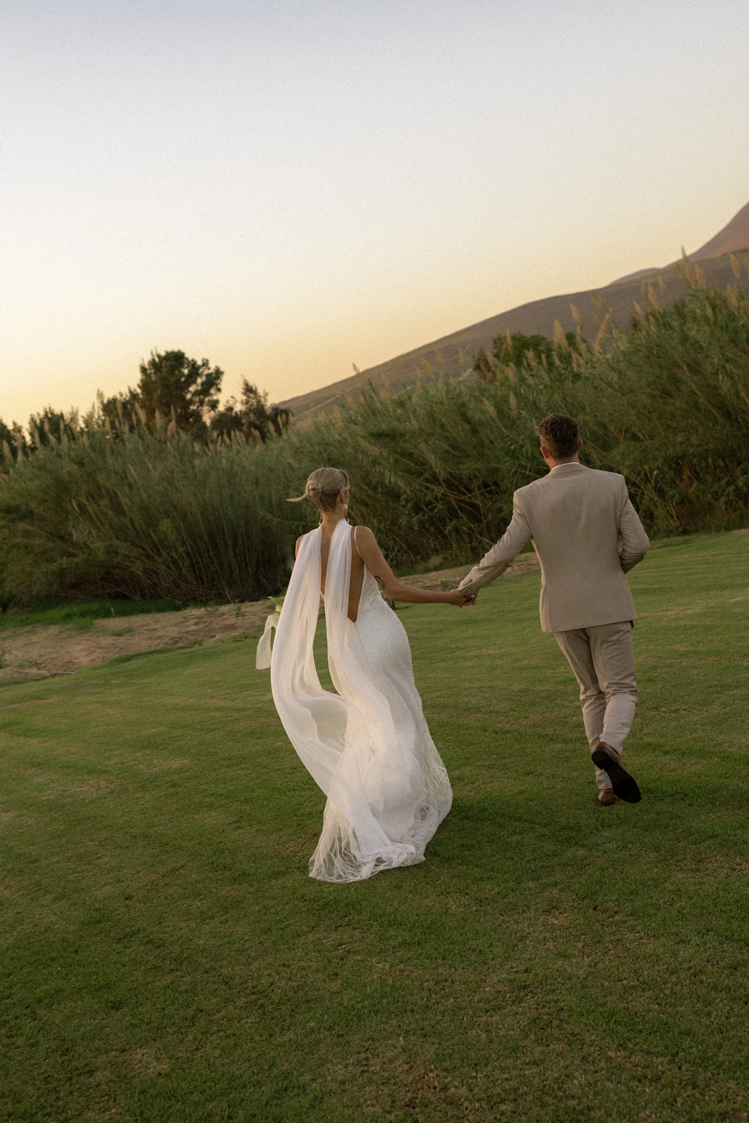 A bride and groom walking hand in hand on a grassy field at sunset, with hills and trees in the background.