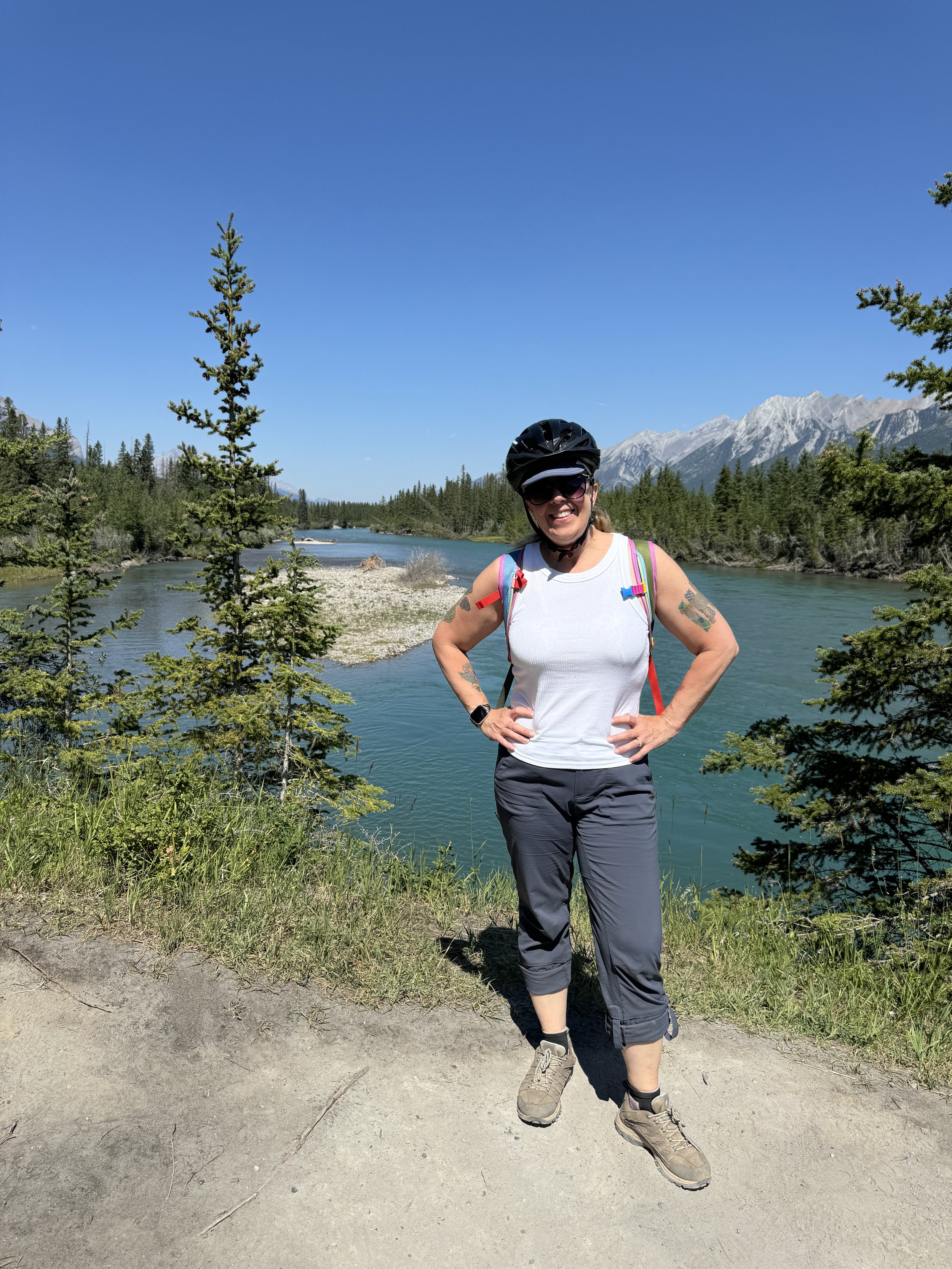 A woman standing outdoors near a river, wearing a helmet, sunglasses, white tank top, and black pants, with mountains and trees in the background on a clear, sunny day.
