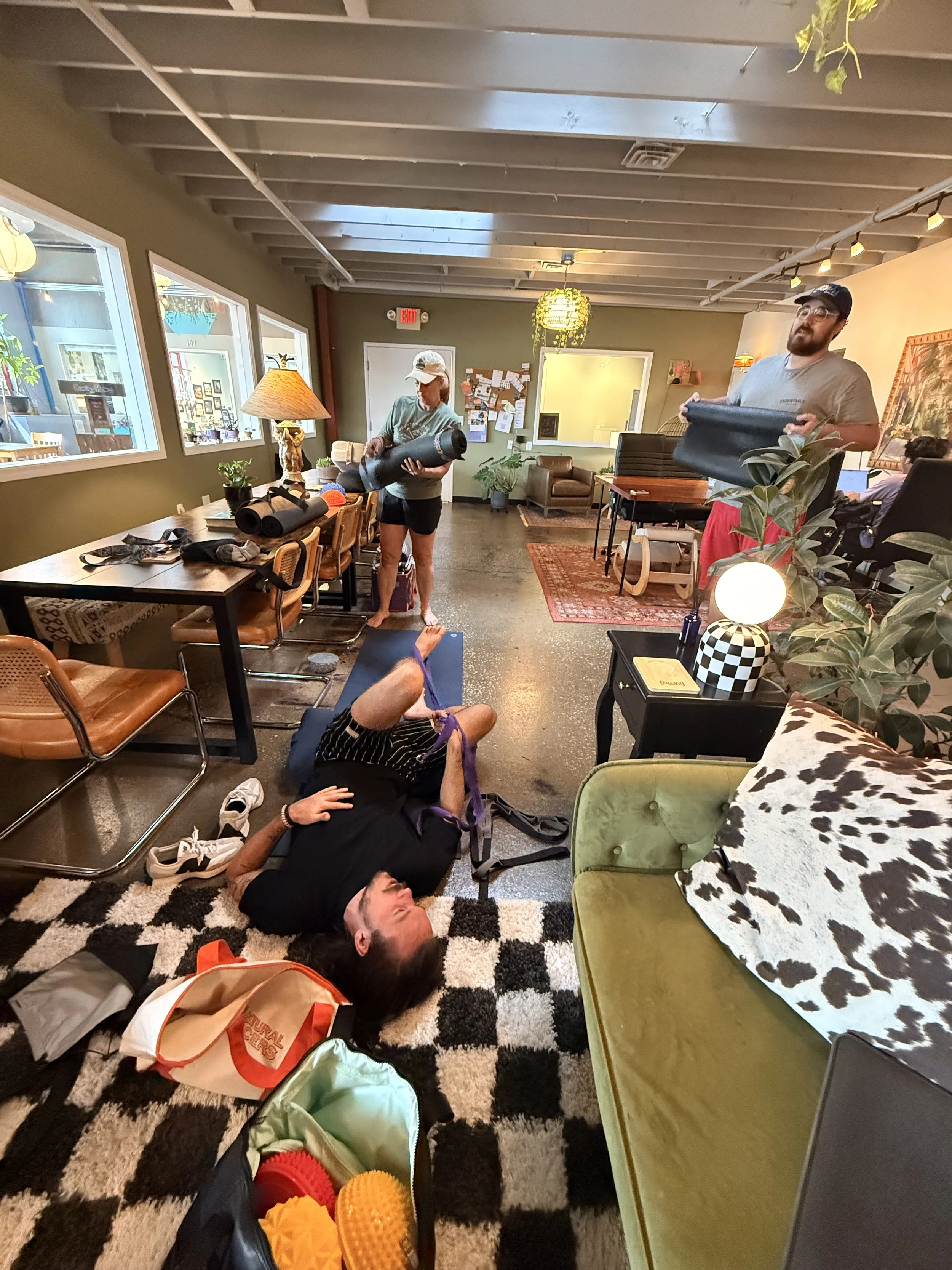A group fitness class in a cozy indoor space with four participants using yoga mats and foam rollers, some lying on the floor and others standing.