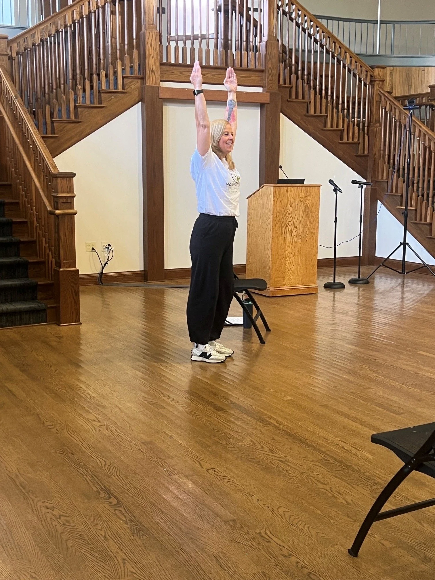 A woman with blonde hair, wearing a white t-shirt and black pants, standing in a room with wooden floors and stairs, raising her arms above her head with a smile.