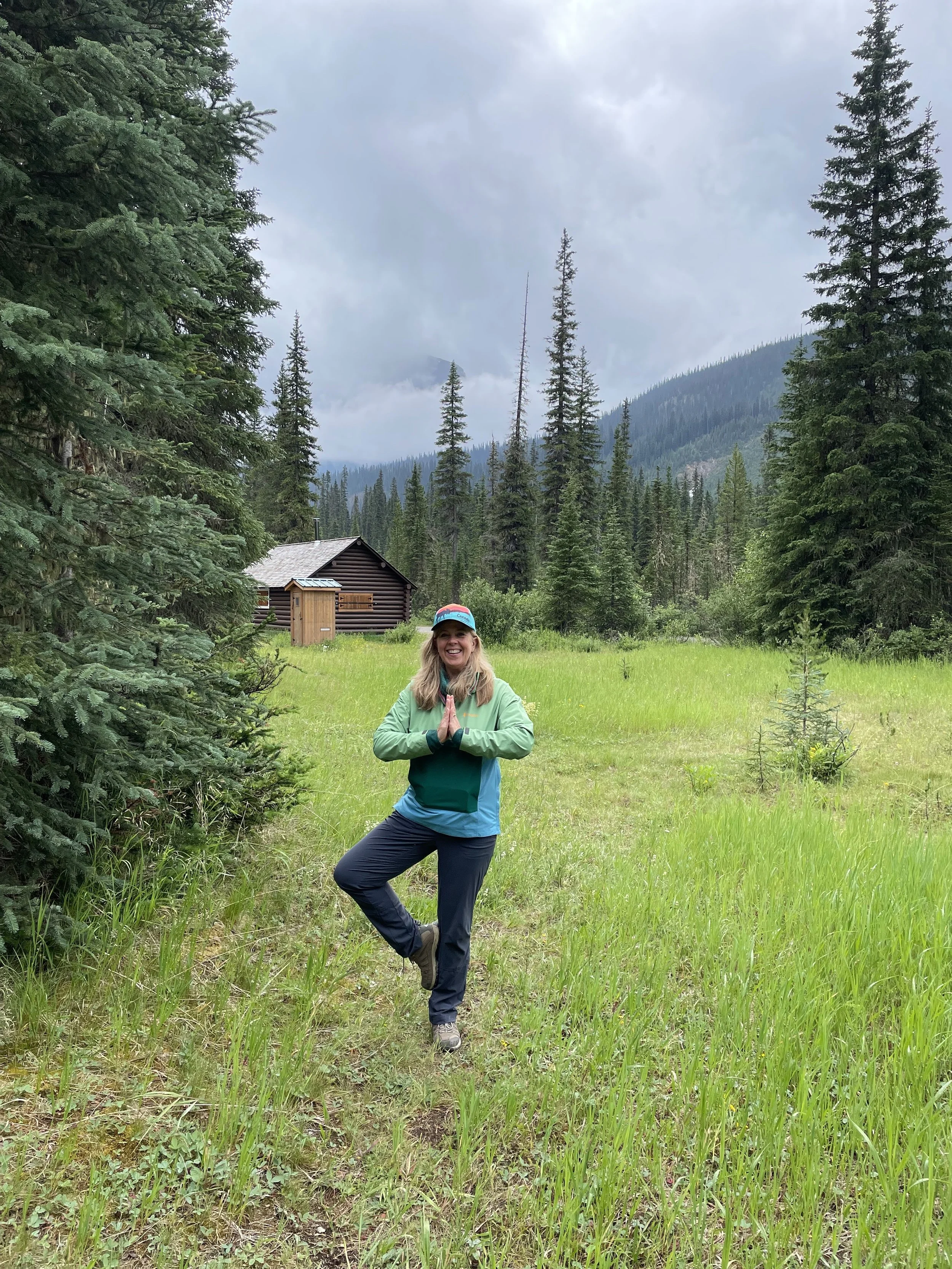 A smiling woman practicing yoga outdoors in a forest clearing, standing on one leg with hands in a prayer position, surrounded by tall evergreen trees and mountainous scenery, under cloudy skies.