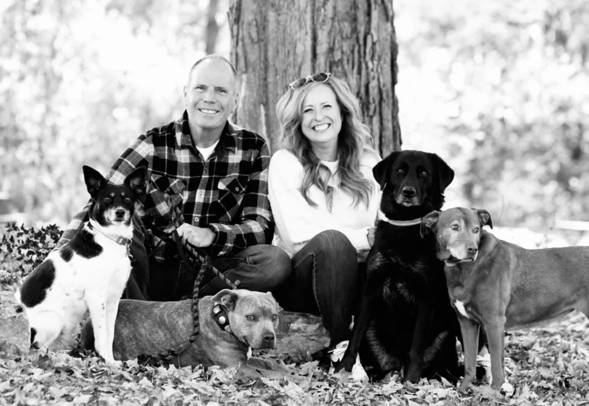 A smiling couple sitting outdoors with four dogs in front of a large tree.