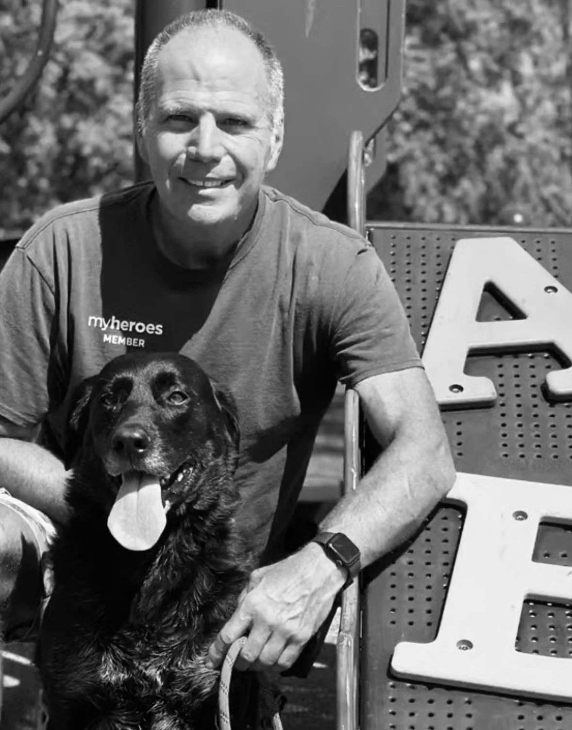 A man smiling outdoors with a dog, both sitting on a bench or structures with a background of trees.