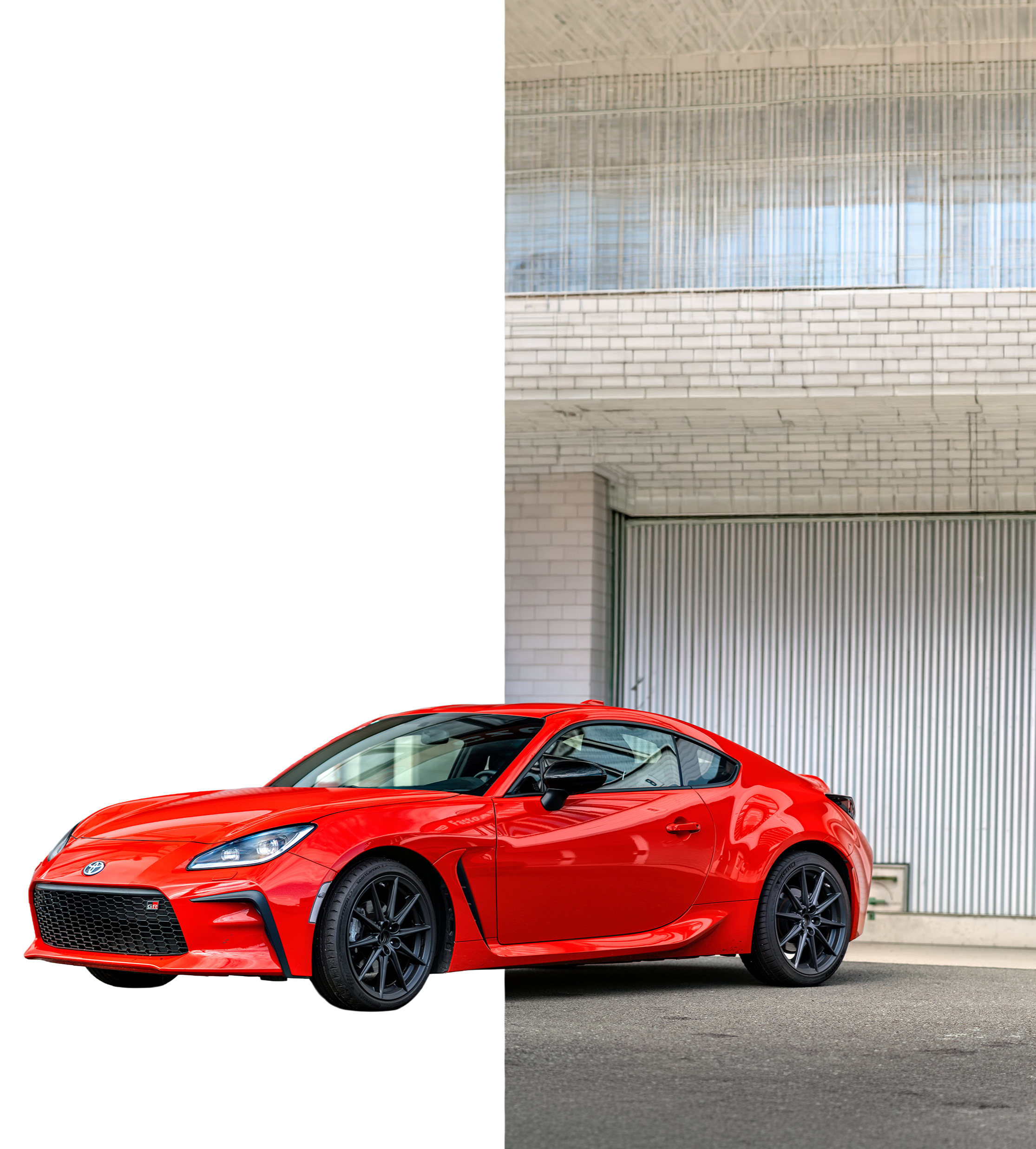Red sports car, likely a Toyota GR 86, parked outside near a modern industrial building with a large garage door and a glass upper floor.