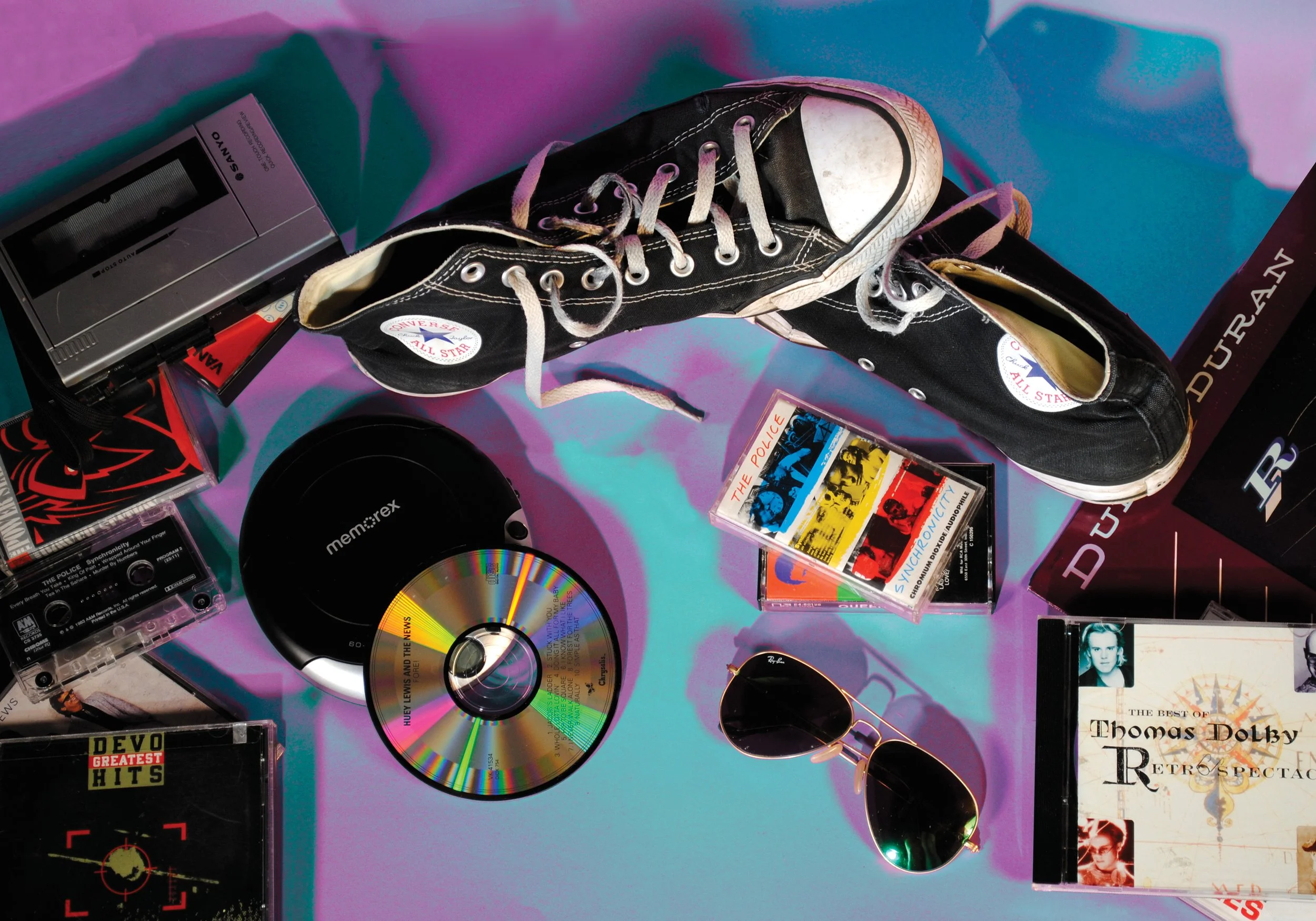 Collection of vintage music and pop culture items including black Converse sneakers, a cassette tape player, CDs, sunglasses, and music albums on a colorful iridescent surface.