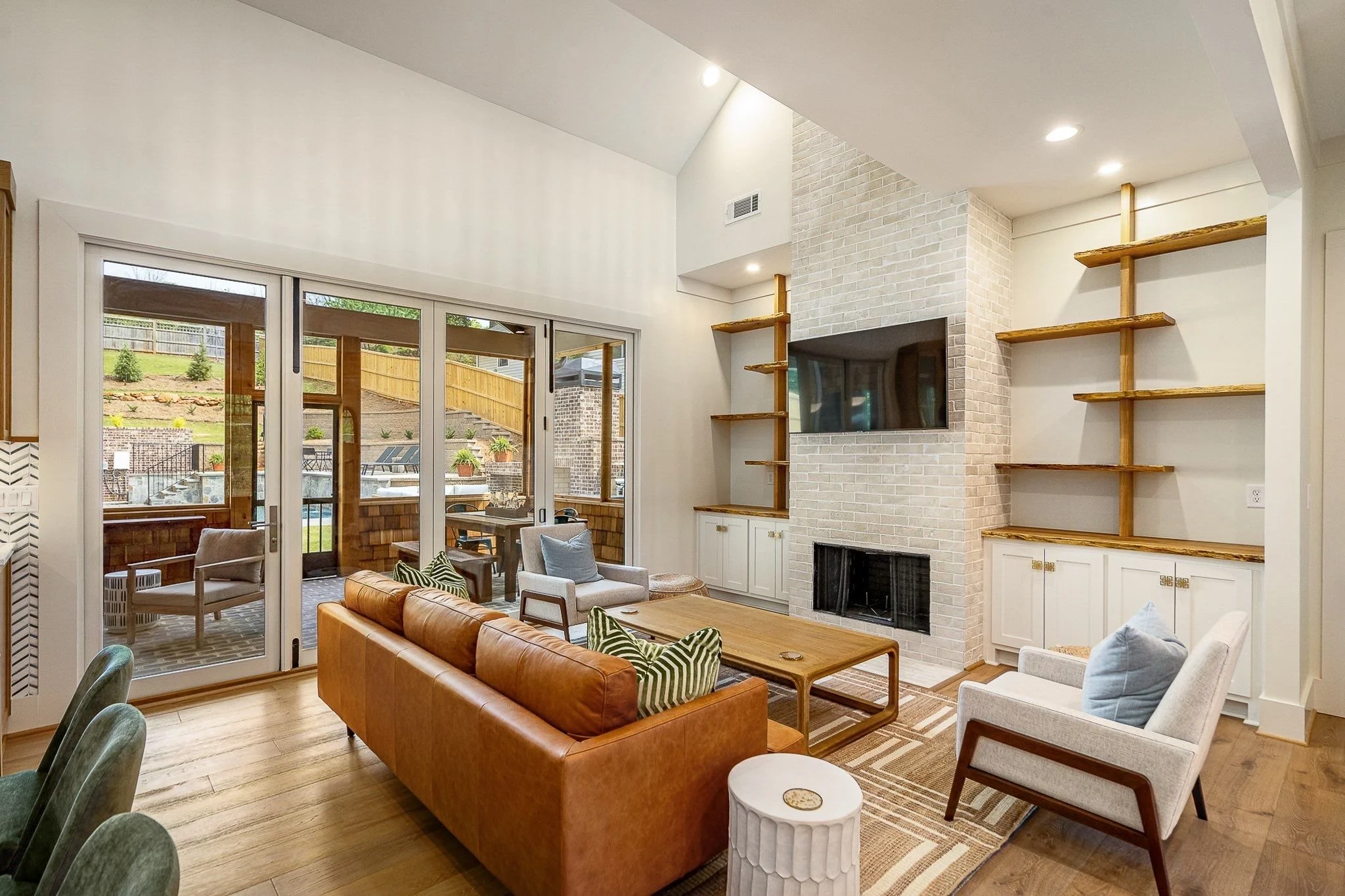 Living room with tan leather sofa, white armchairs, wooden coffee table, and a fireplace with built-in shelves. Large sliding glass door opens to an outdoor patio with outdoor seating and a grassy hill.