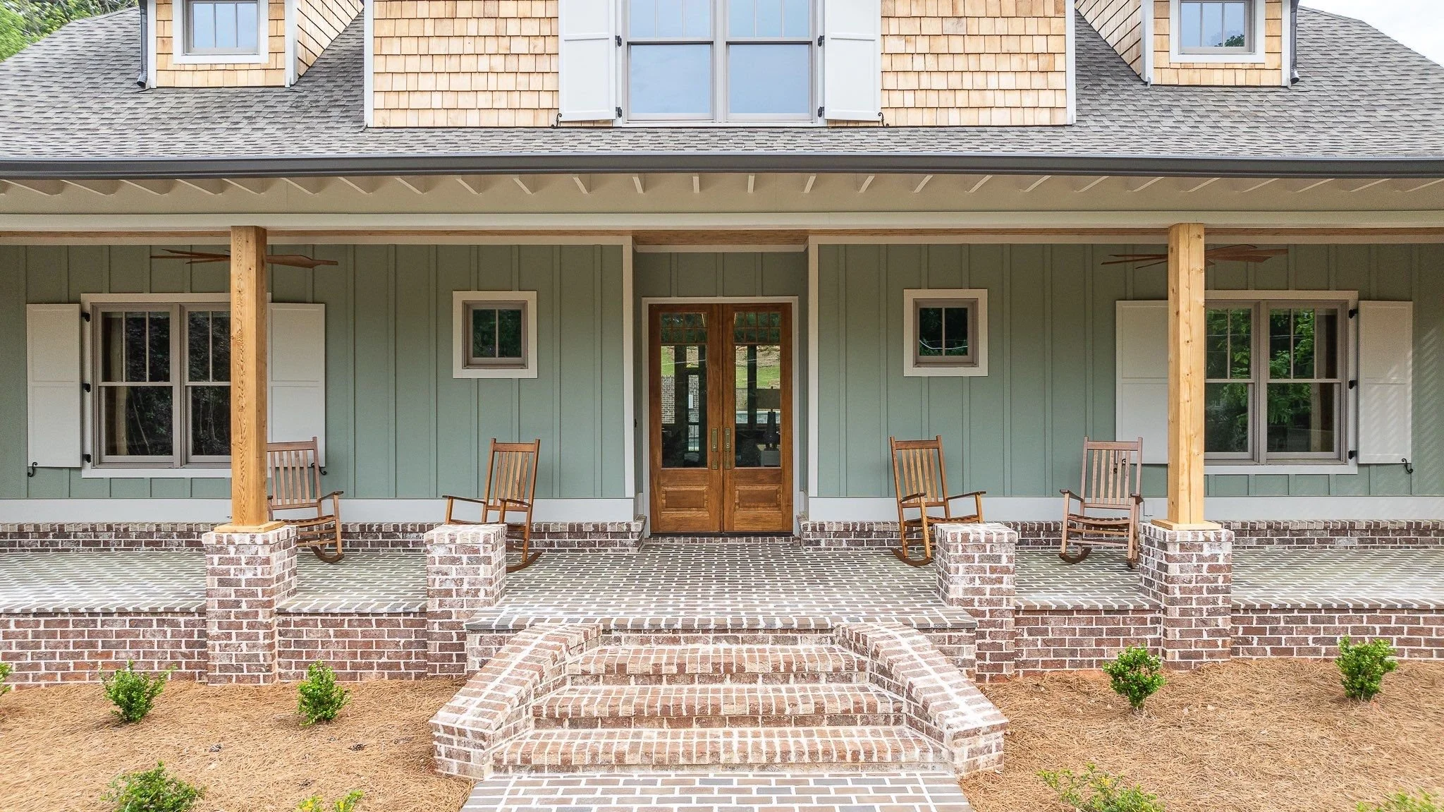 Front porch of a house with brick stairs, green exterior walls, two rocking chairs, and wooden double doors.