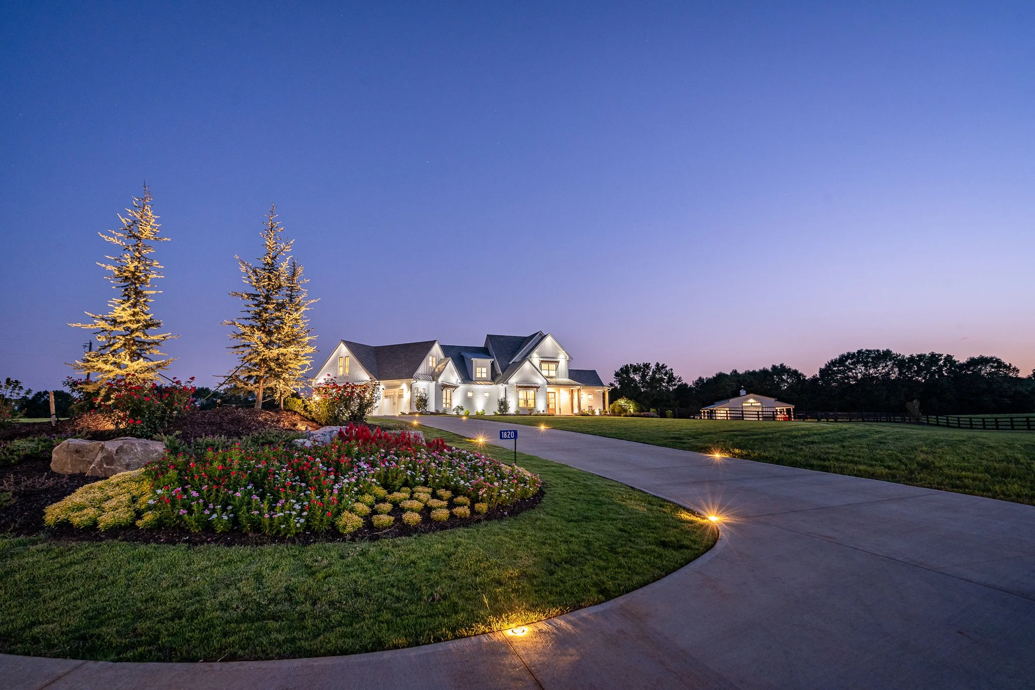 A large white house with a dark roof, illuminated at dusk, surrounded by a landscaped yard with colorful flowers, trees, and pathway lights.