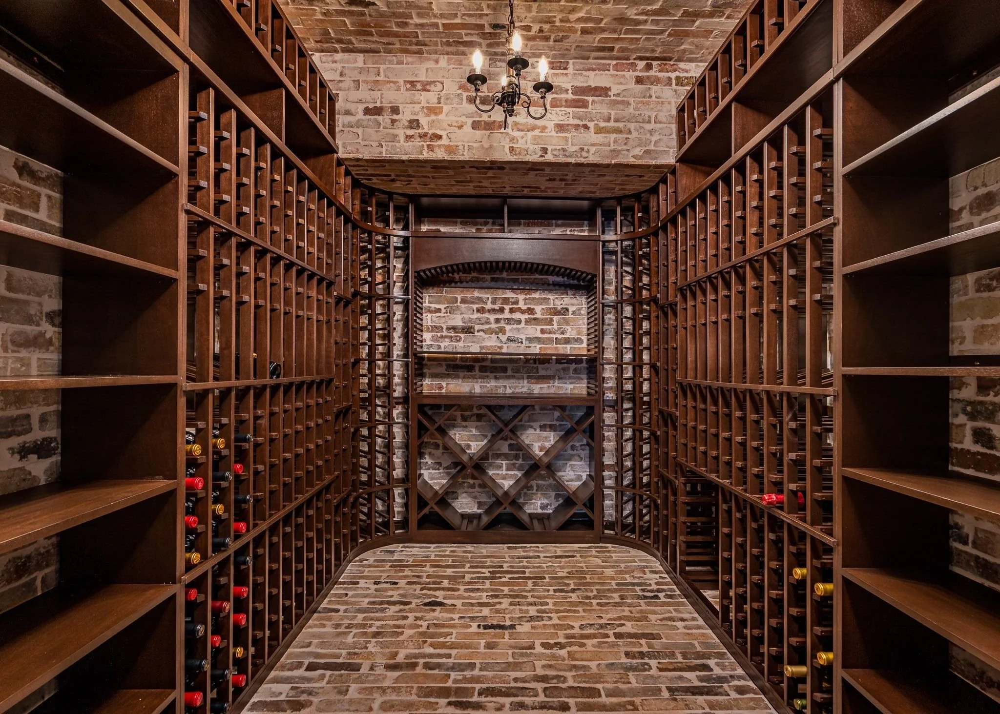 A wine cellar with brick walls, wooden shelves, and wine bottles stored horizontally. There is a central rack and a chandelier hanging from the ceiling.
