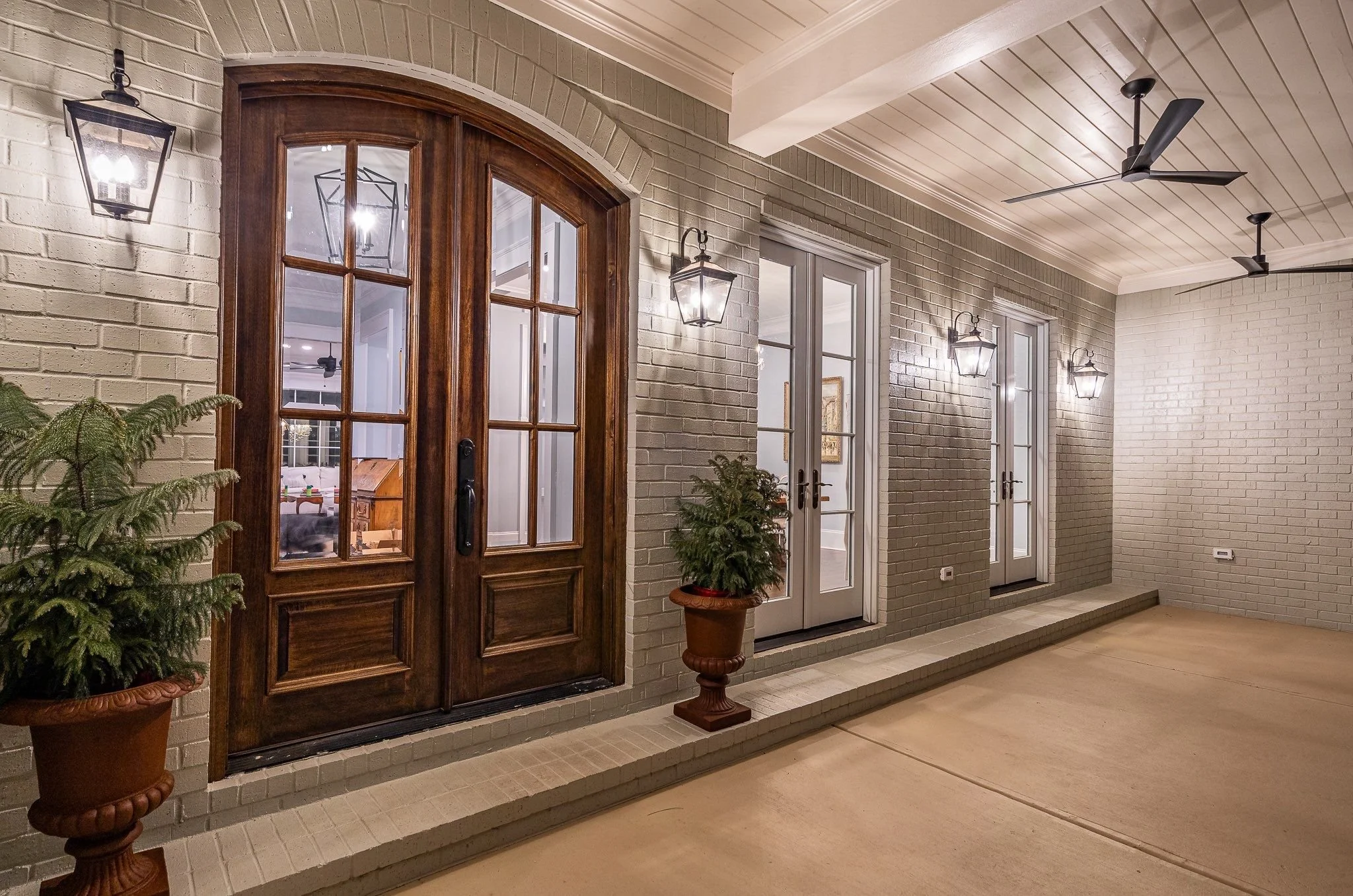 Patio area with gray brick wall, white ceiling, ceiling fans, outdoor wall lanterns, wooden and white-framed glass doors, potted plants, and a concrete floor.