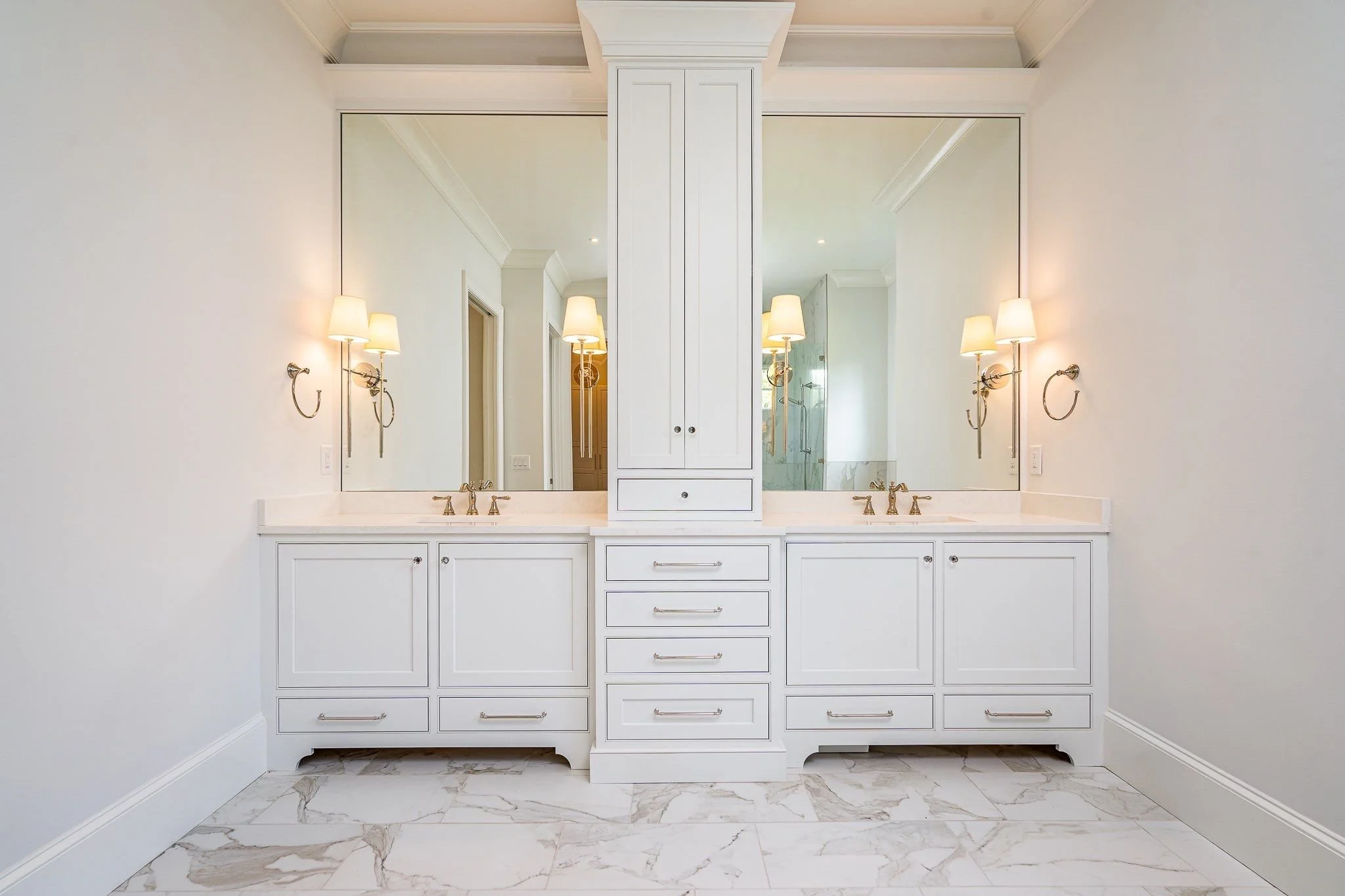 Double vanity with two mirrors, white cabinetry, marble countertops, wall-mounted sconces, and a large central cabinet, in a bright bathroom with marble tile flooring.