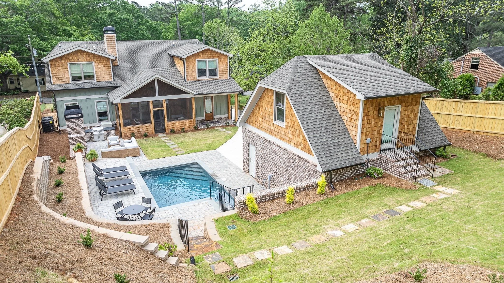 A backyard with a swimming pool, patio furniture, a screened porch, and two houses with wood and brick exteriors, surrounded by a wooden fence and green trees.