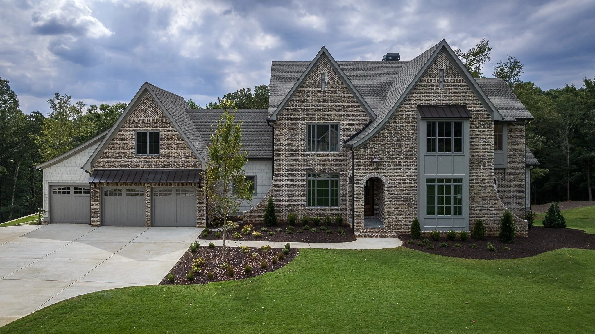 Large brick and siding house with front yard, green lawn, concrete driveway, trees, and a cloudy sky.