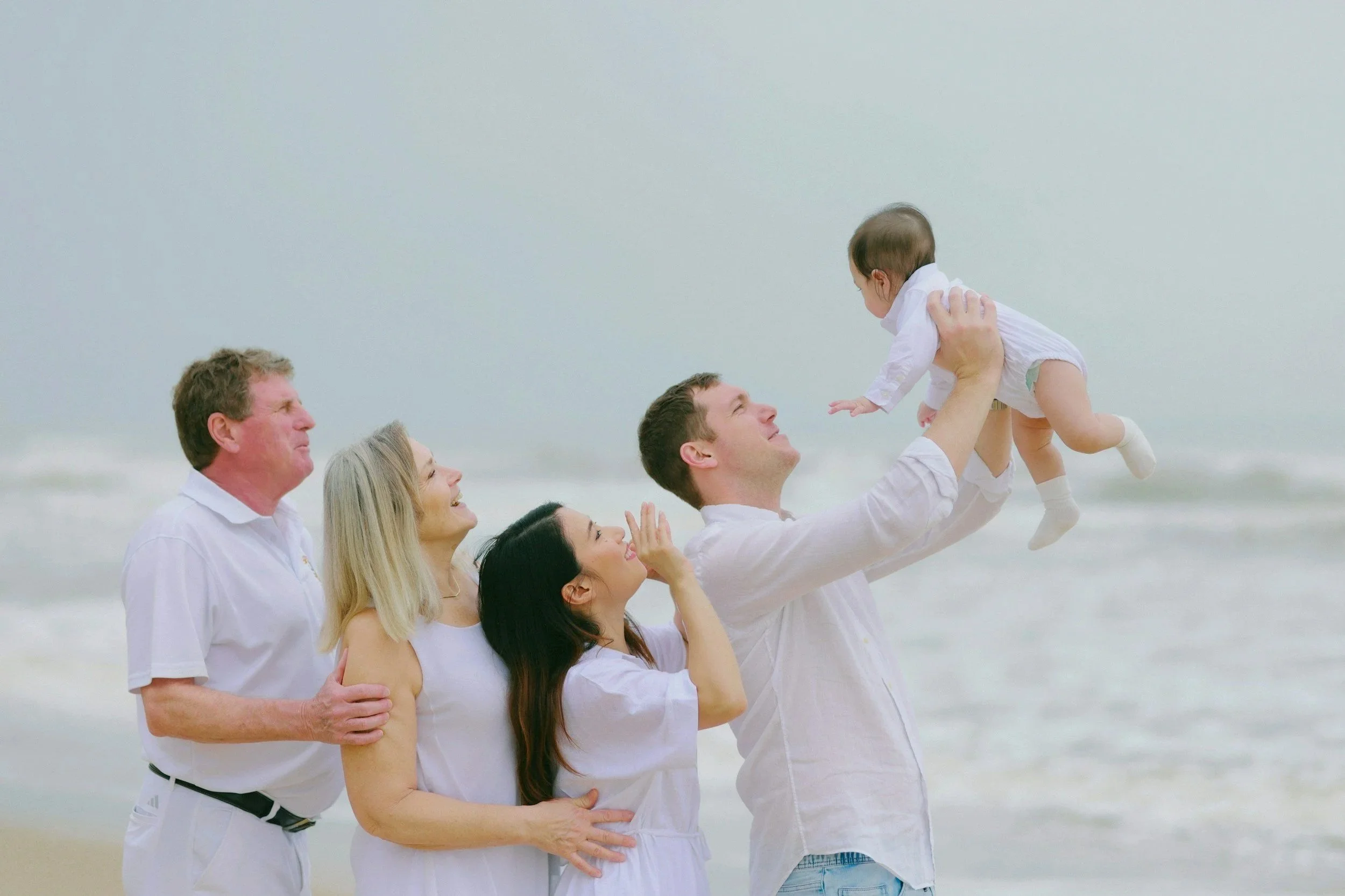 Family groups of grandparents, parents, and children enjoying at the beach, with one man lifting a baby girl into the air.