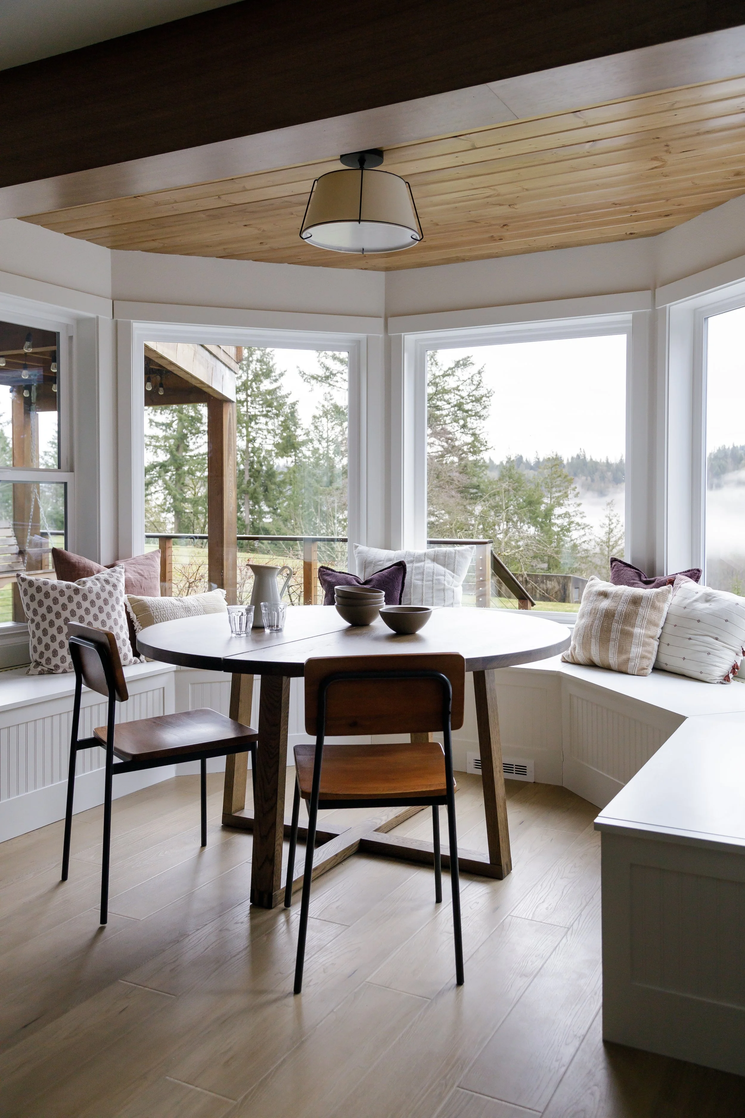 Bright dining nook with a round table, three chairs, and window seating with pillows, overlooking a scenic outdoor view of trees and fog.