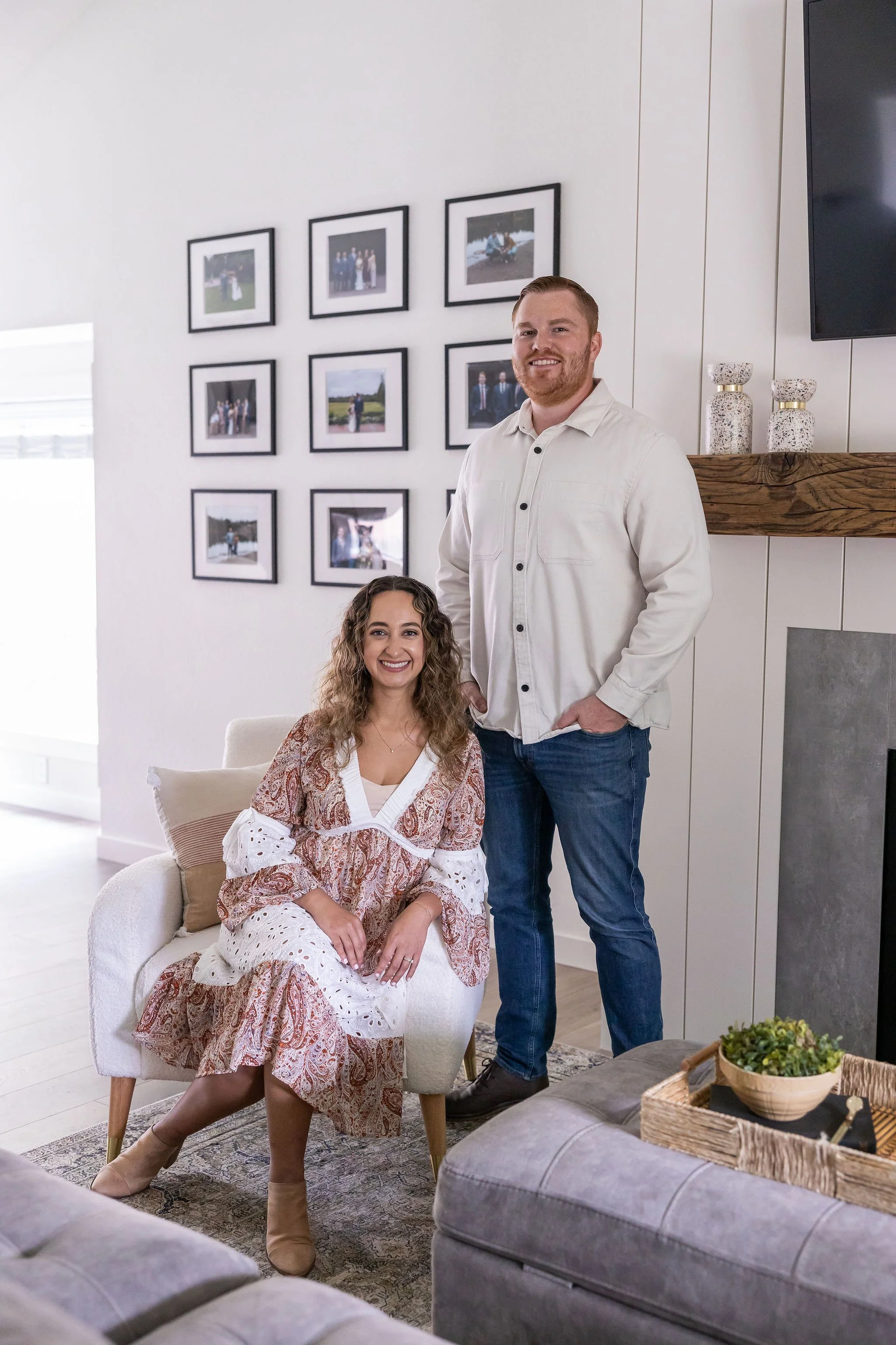 A smiling woman sitting on a cream-colored armchair and a smiling man standing beside her in a modern living room with framed photos on the wall.