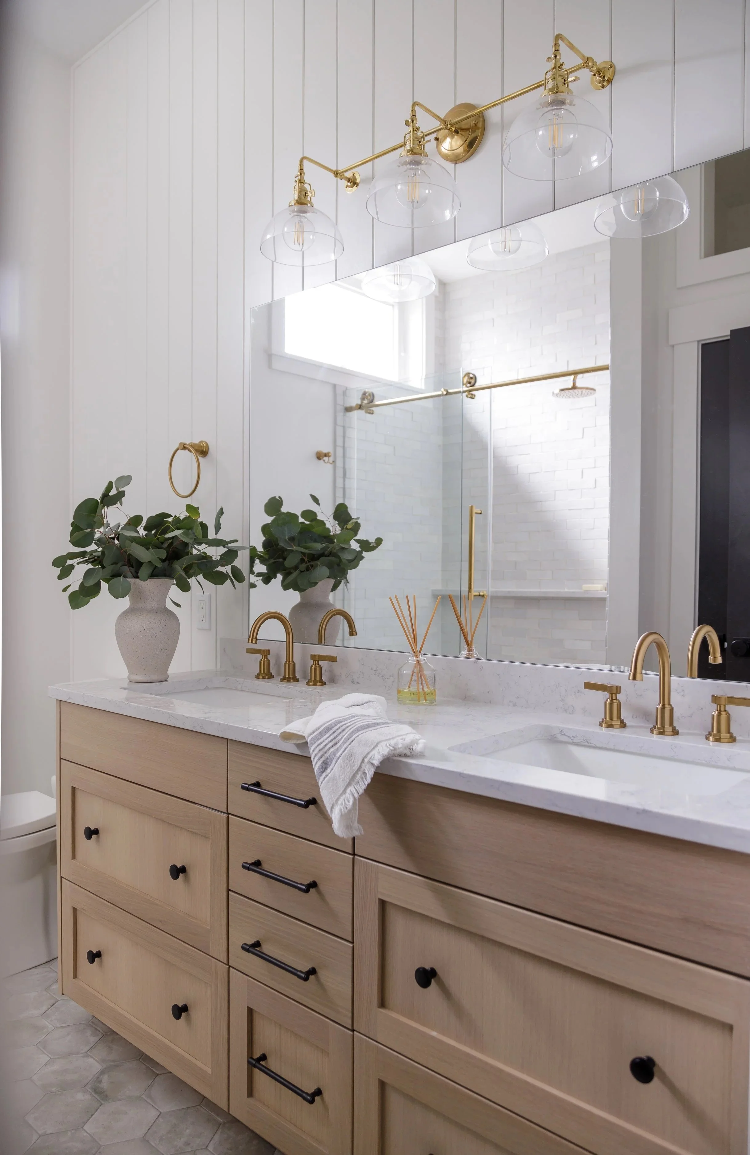 Modern bathroom with light wood vanity, marble countertop, gold fixtures, large mirror, white tile walls, and decorative plants.