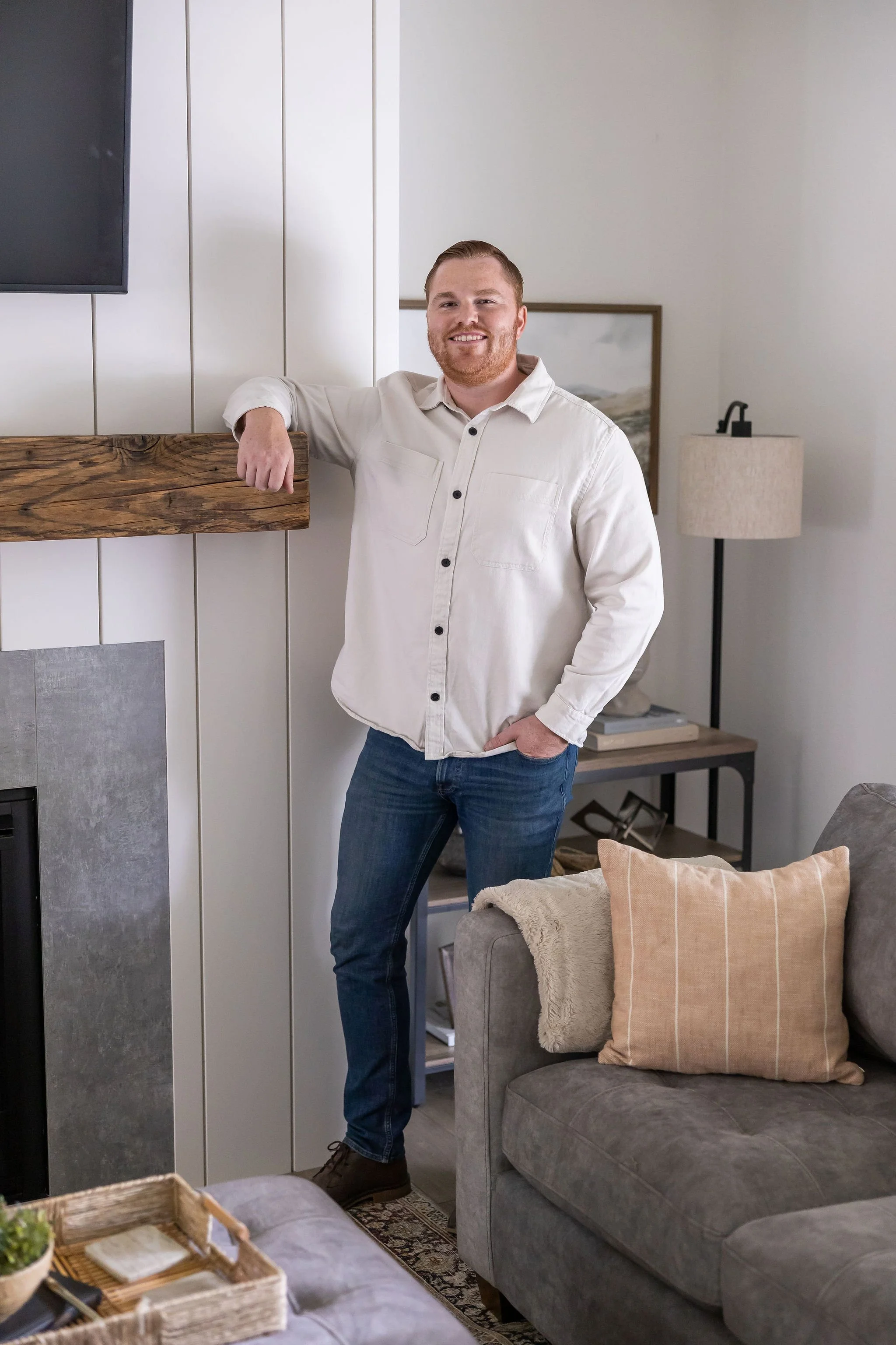 A man with a beard and smiling, standing in a living room, leaning on a wooden fireplace mantle, wearing a white shirt and blue jeans.