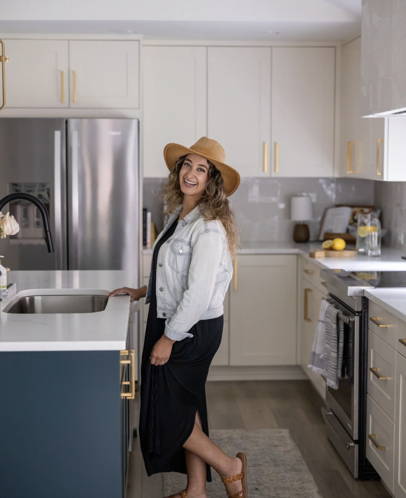 A woman with curly hair wearing a straw hat, white denim jacket, and black dress standing in a modern kitchen, smiling at the camera.